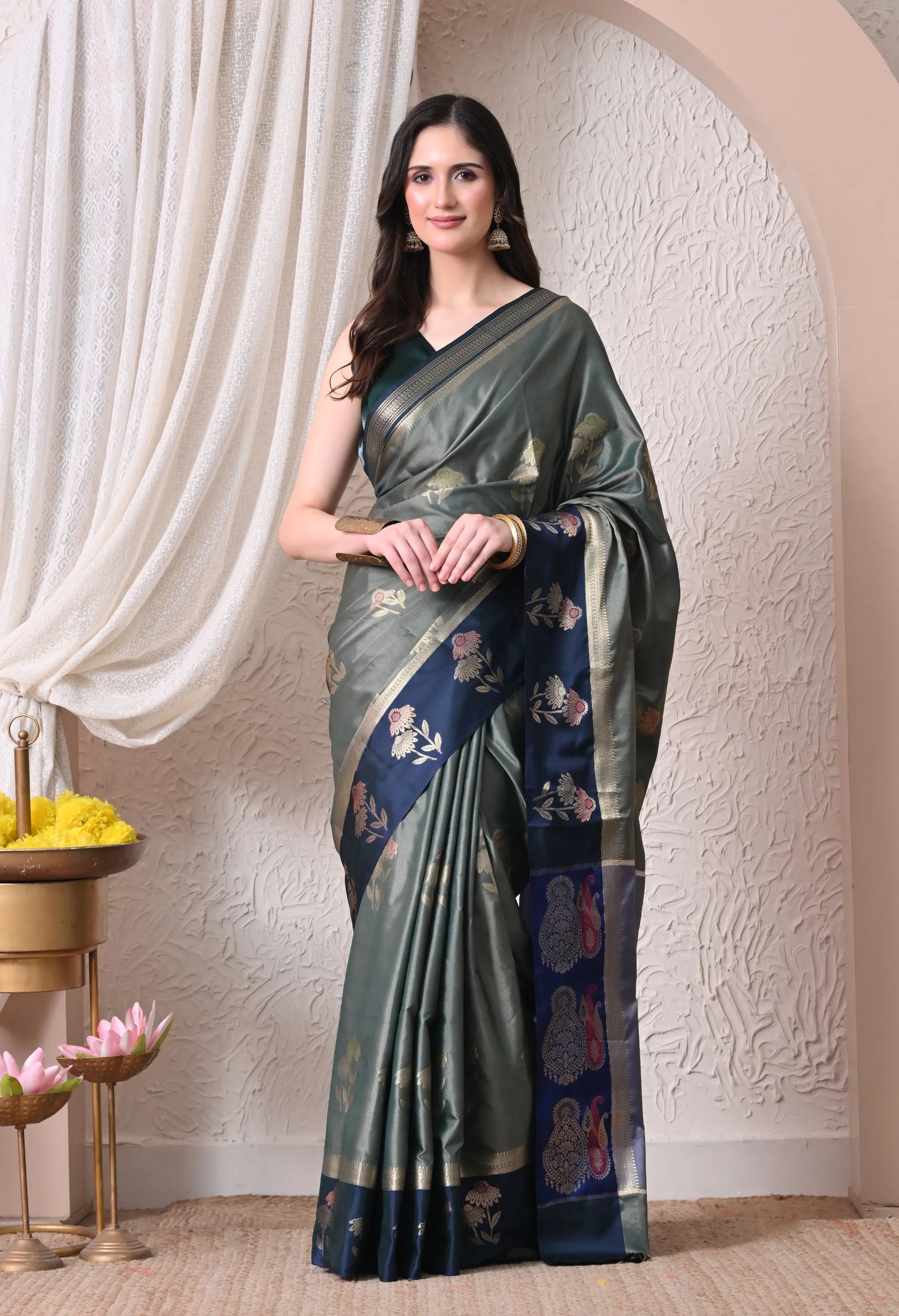A woman modeling an olive grey silk blend saree featuring scattered floral motifs and a contrasting deep navy blue border and pallu intricately woven with gold-toned patterns.
