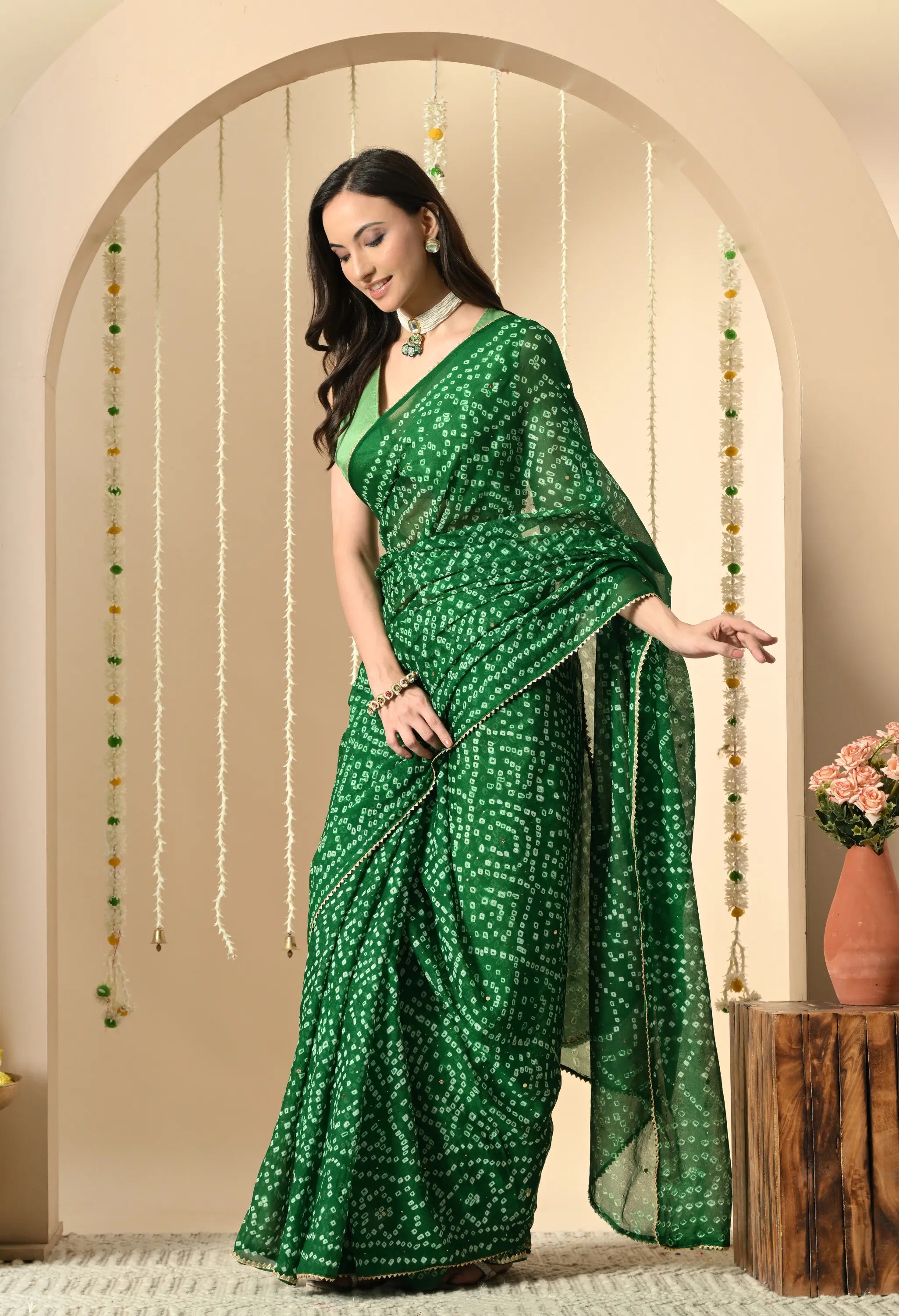 A woman is pictured in a side-profile view, looking down and smiling while wearing a vibrant green georgette sari from The Weavers Shop by Raavi. The sari features a traditional handcrafted white Bandhani tie-dye pattern and a delicate gold border. She is styled with a sleeveless light green blouse and a pearl choker, standing against a neutral-toned archway decorated with floral garlands.