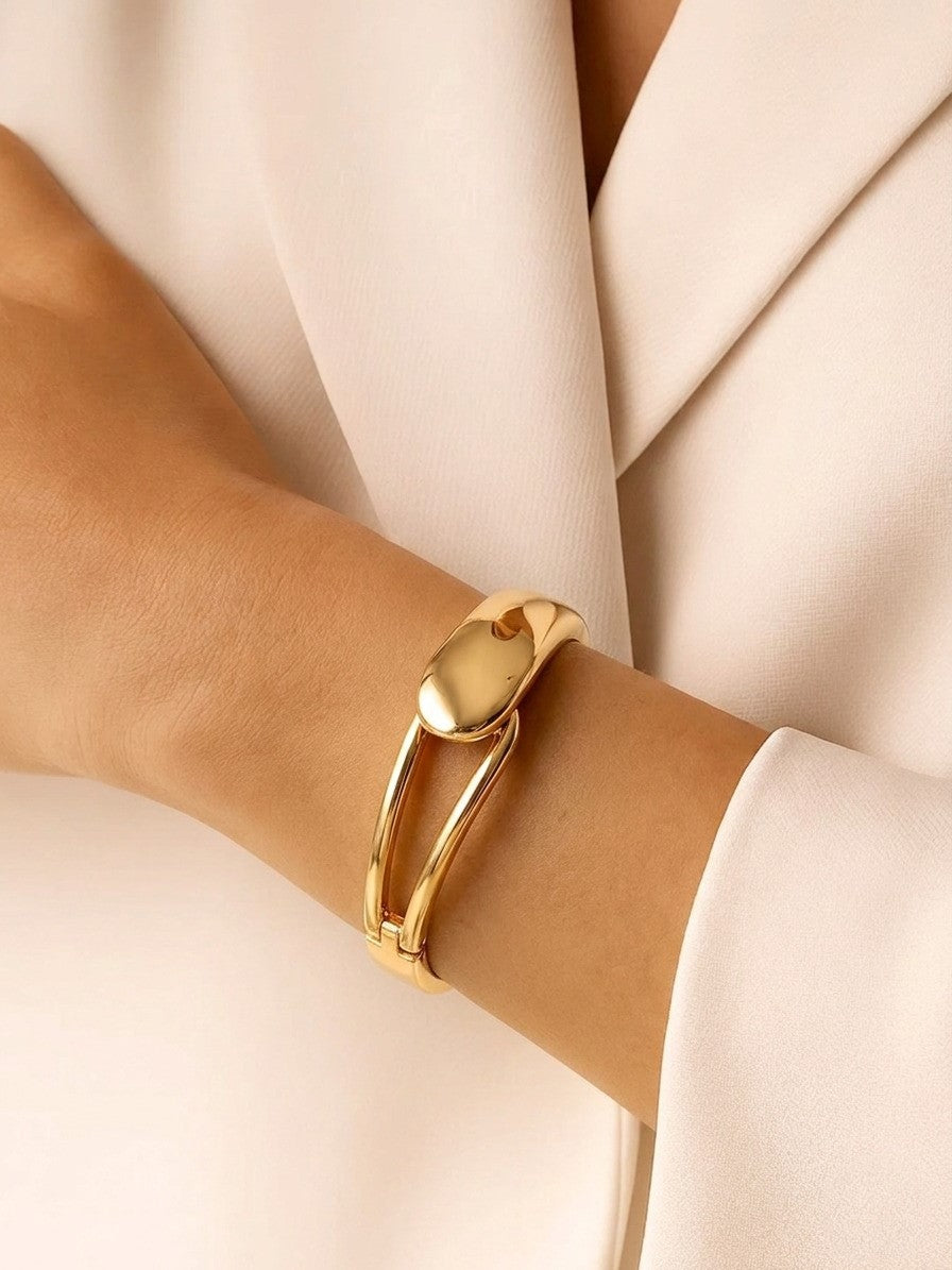 A person wearing a thick, polished gold statement bangle with a large domed oval center on their wrist.
