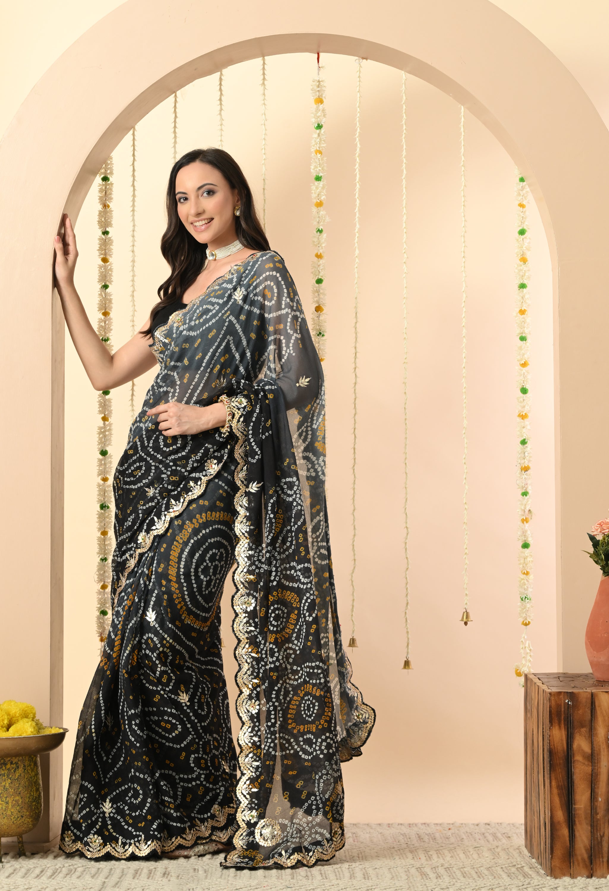 A woman stands leaning against a cream-colored archway, showcasing a sophisticated black georgette saree from The Weavers Shop by Raavi. This handcrafted Rajasthani Bandhani saree, designed for a festive event or high-end celebration, features intricate white and yellow tie-dye patterns and is highlighted by a traditional gold Gota Patti scalloped border. She is styled with a sleeveless black blouse and a white pearl choker, posed in a setting decorated with jasmine garlands.