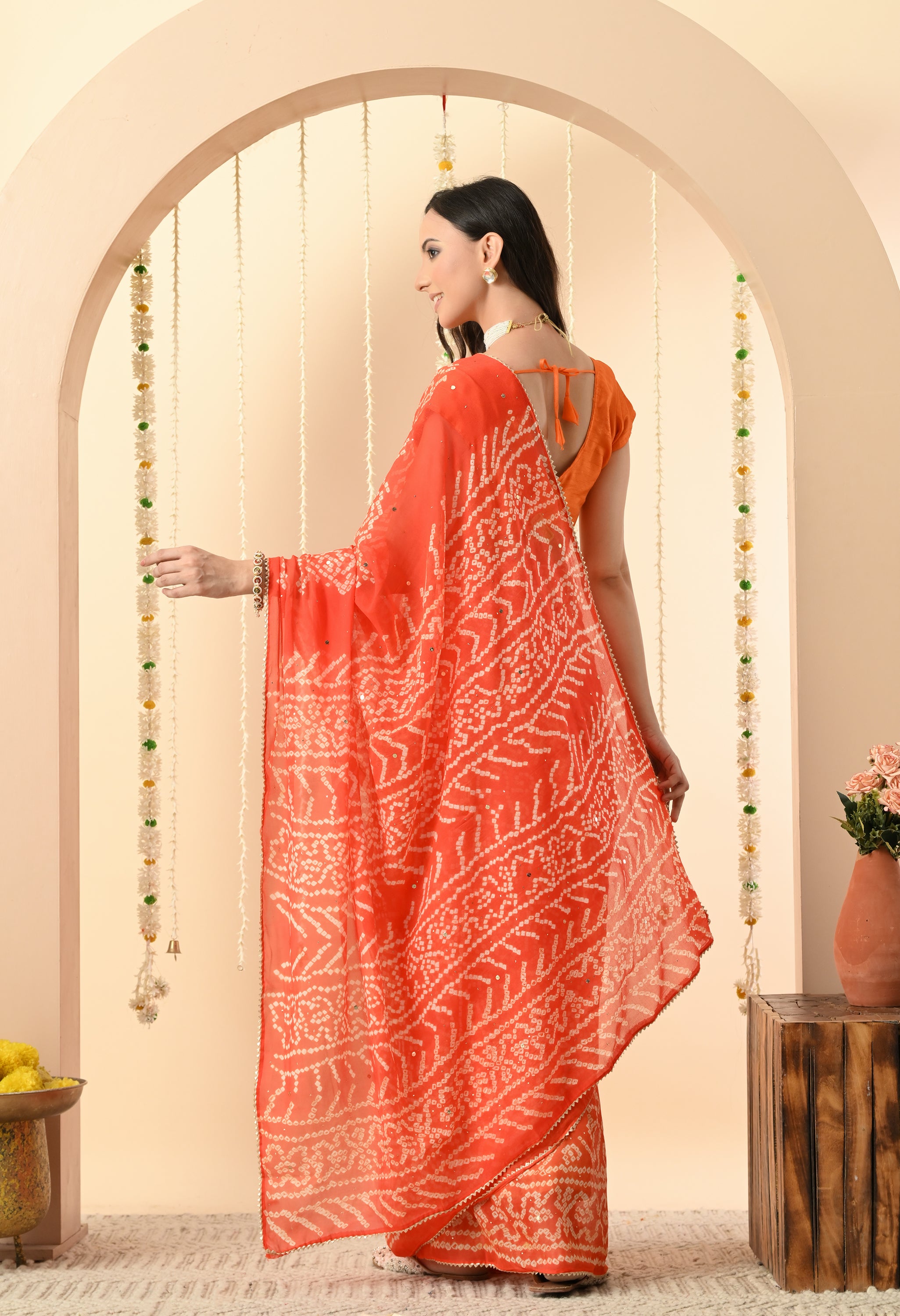 A woman is pictured from the back, showcasing the full drape of an original handmade orange Bandhej saree from The Weavers Shop by Raavi. The georgette fabric displays a complex tie-dye design, ideal for festive occasions. The view highlights the back of the matching orange blouse and the elegant flow of the saree.
