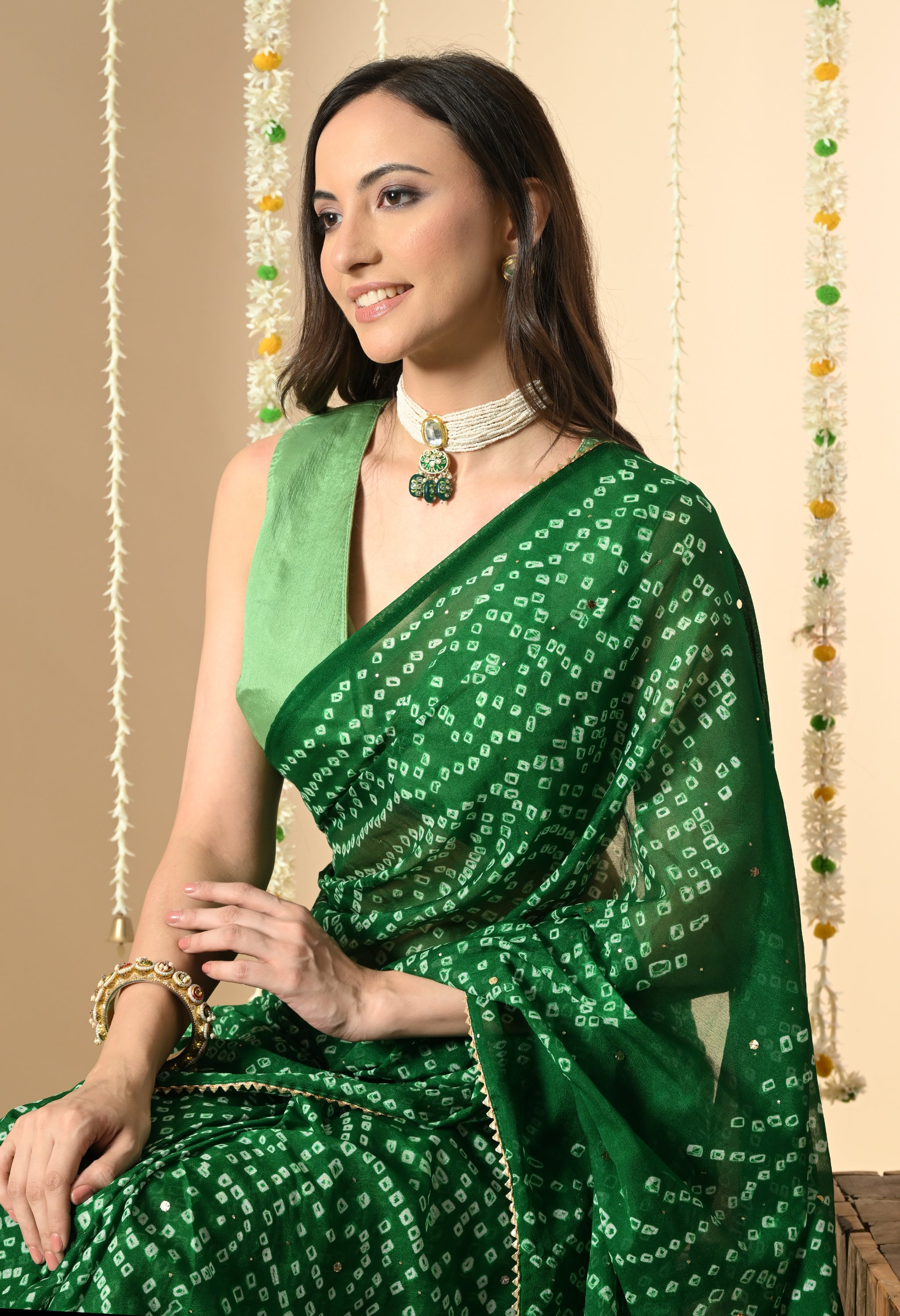 A woman smiles while looking down, seated in a stunning green georgette Bandhej sari from The Weavers Shop by Raavi. This festive look is completed with traditional jewelry and a soft, elegant drape, ideal for an event like Navratri or a wedding.