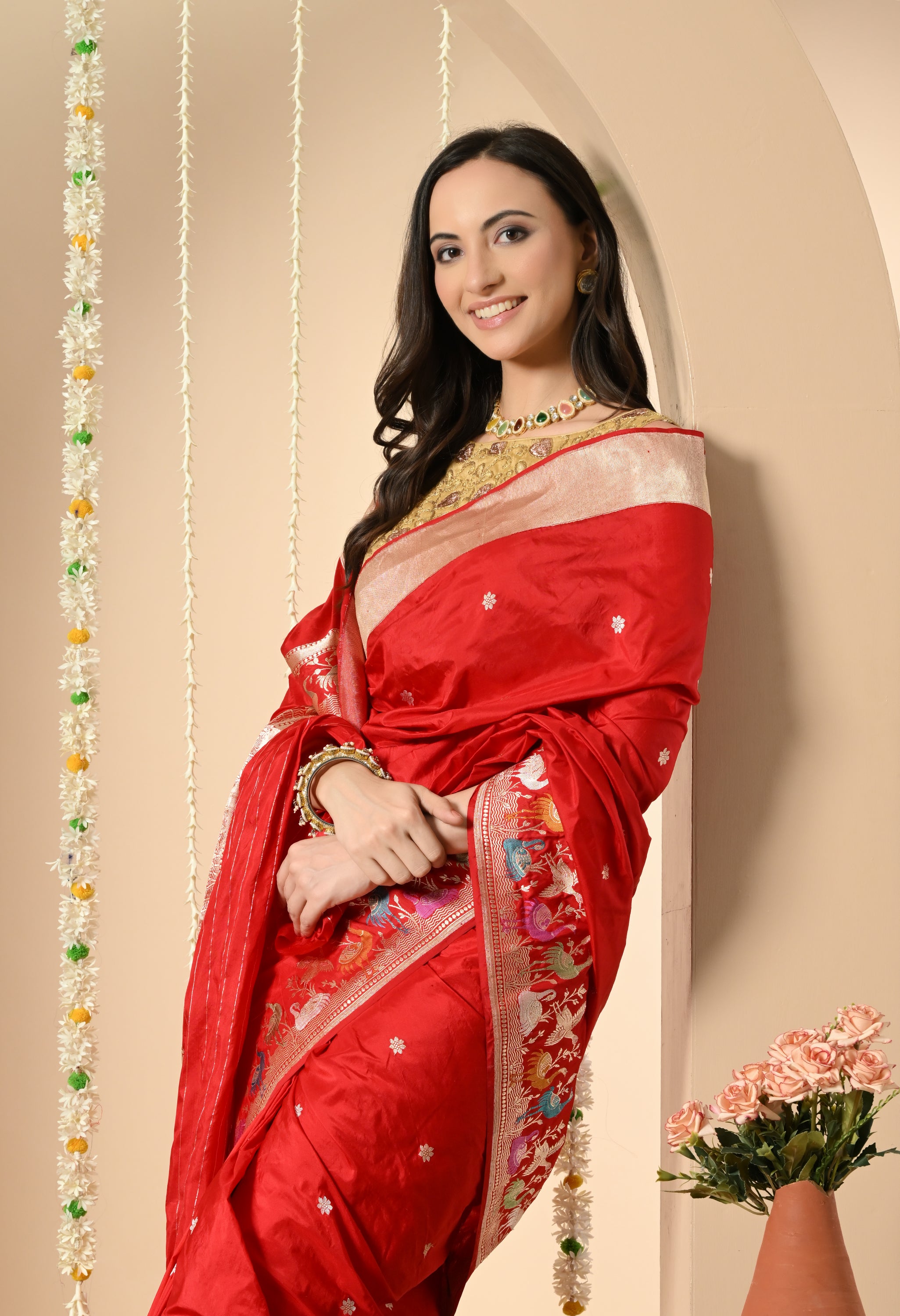 A Zoom-in view of  A beautiful woman modelling a luxurious vibrant red Katan silk saree from The Weavers Shop by Raavi (Silk Mark certified). The image highlights the saree's detail: subtle woven Zari motifs (butis) on the body and the wide, multicoloured Shikarga border with intricate Zari work depicting animals and figures. She is wearing a gold blouse and traditional jewellery.