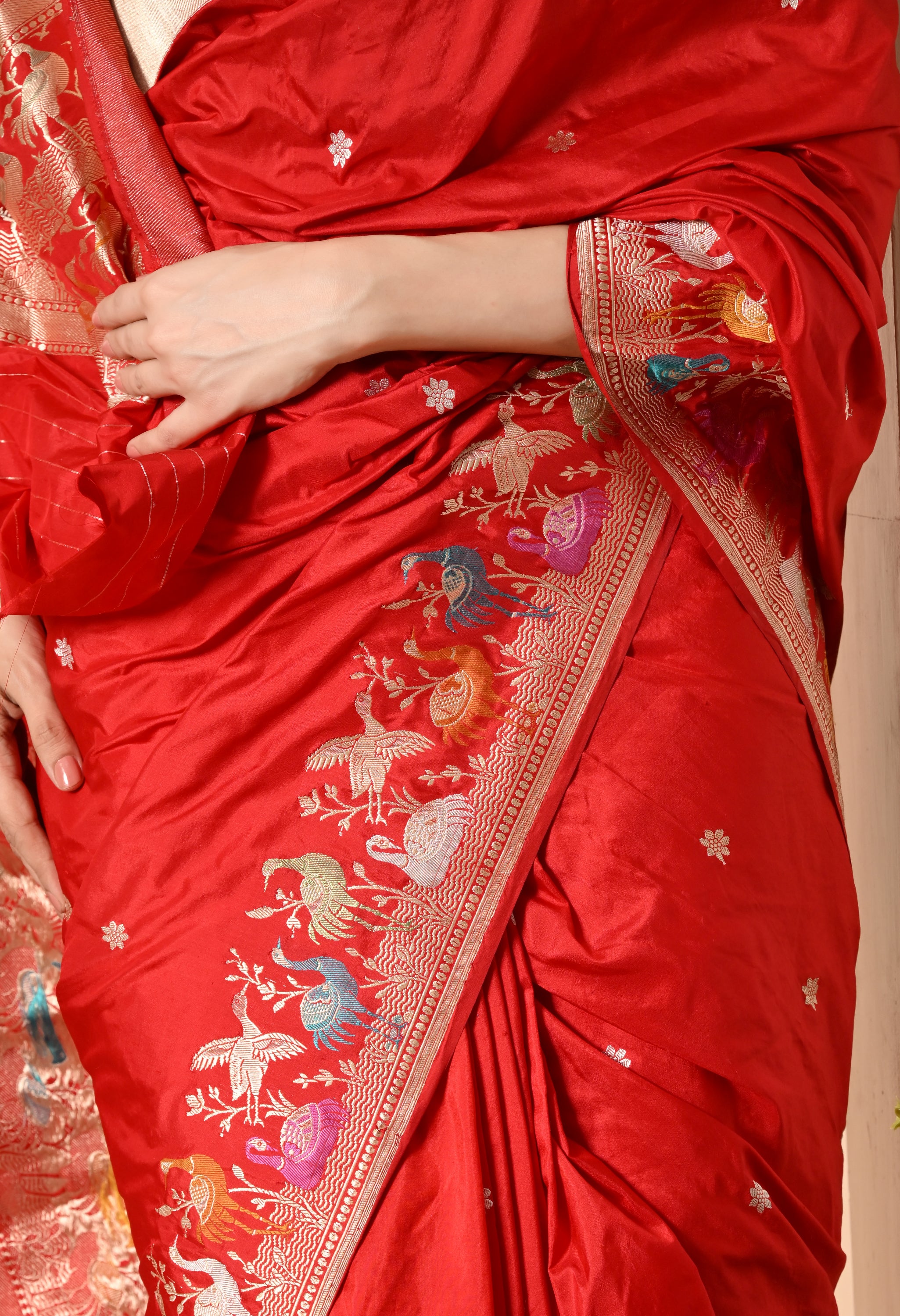 Zoom-in view showcasing the product detail: A close-up of a luxurious vibrant red Katan silk saree from The Weavers Shop by Raavi (Silk Mark certified). The image highlights the Zari motifs (butis) on the fabric and the intricate, multicolored Shikarga border. The border features detailed Zari work with woven motifs of birds and other animals. A woman's hands lightly touch the draped fabric.