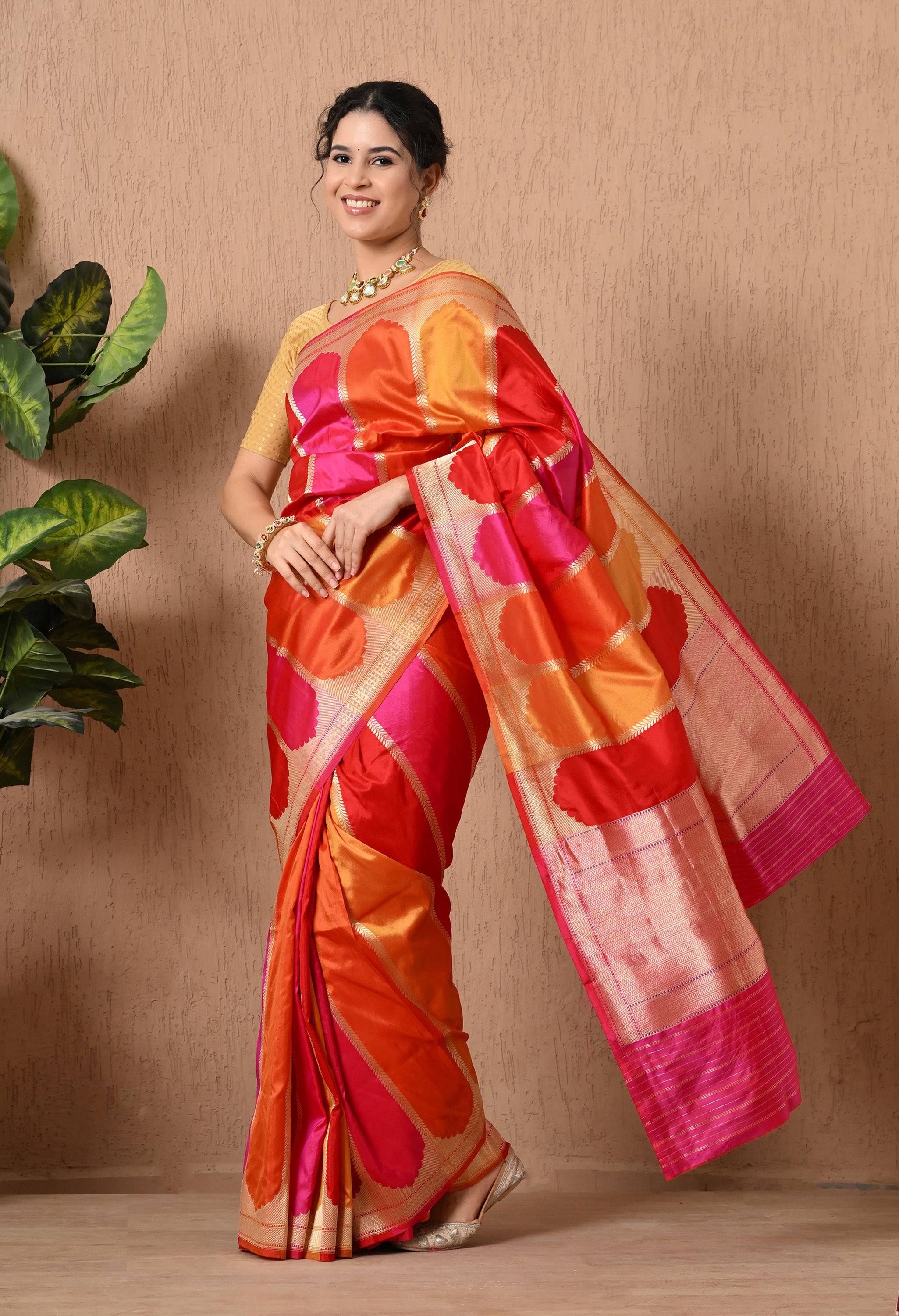 A woman stands in a profile view, showcasing a vibrant Pure Katan Silk saree from The Weavers Shop by Raavi. This authentic Banarasi garment features the traditional Rangkat weaving technique with bold diagonal blocks of color in shades of pink, orange, and red. The saree is adorned with intricate Zari work, including a shimmering gold border and decorative patterns. This Silk Mark Certified piece is styled with a gold blouse and a statement necklace.