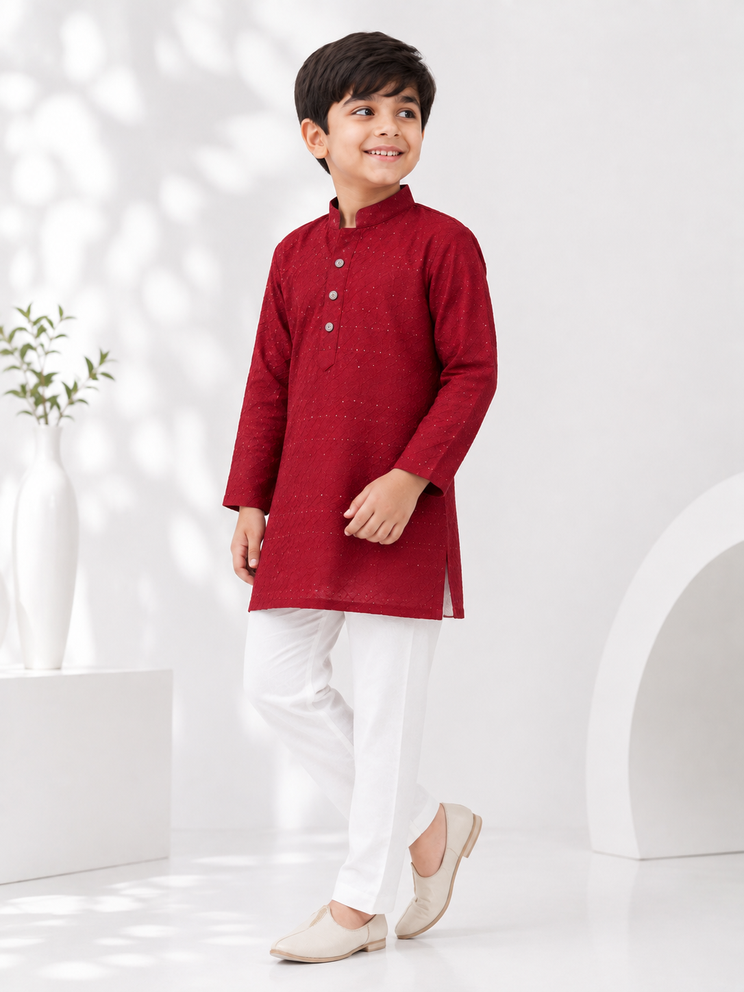 "Young boy wearing a maroon full-sleeve embroidered kurta pajama set with white pajama trousers and beige traditional jutti shoes, posing in a bright white studio with a modern minimalist background.
