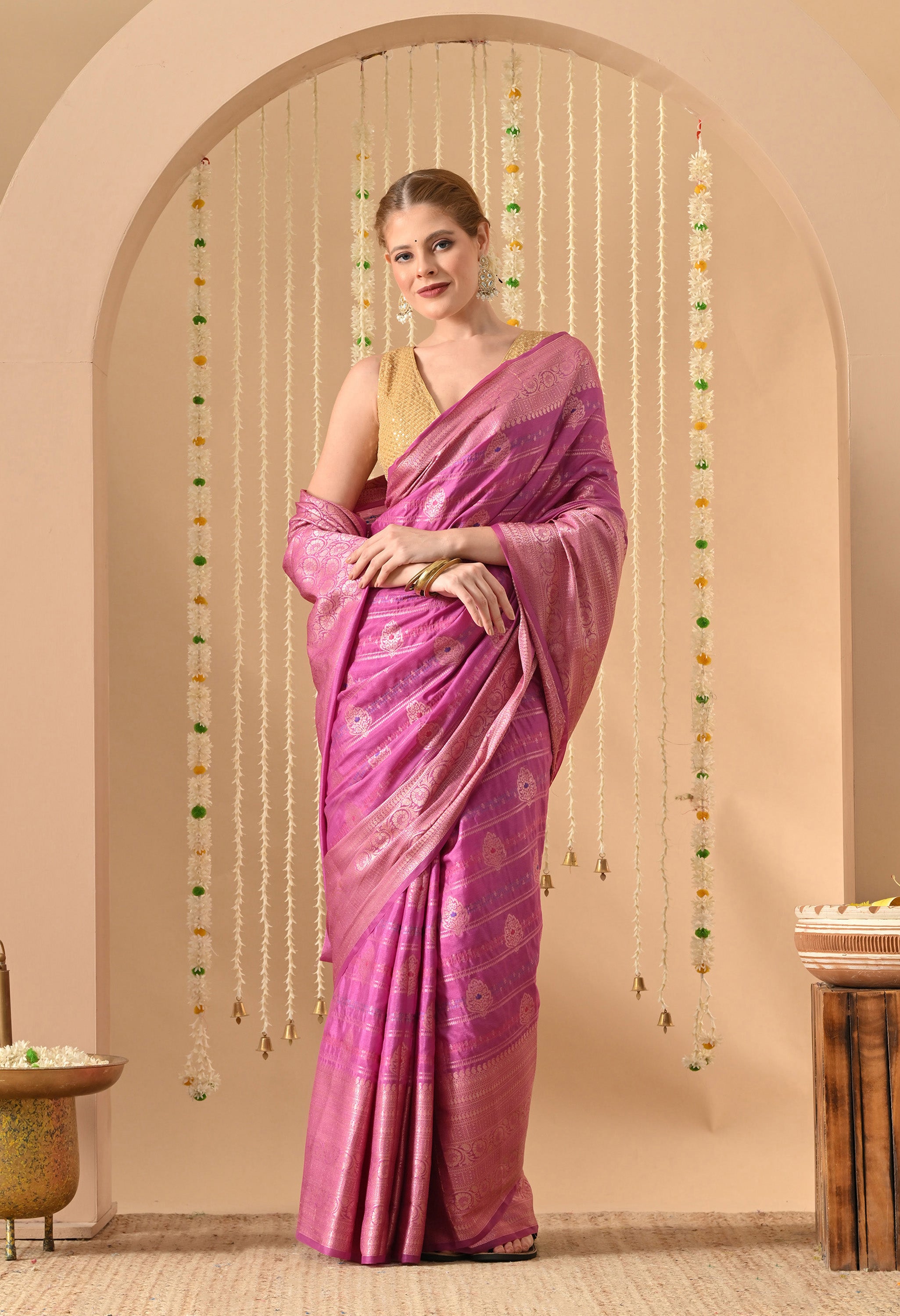 A model poses in a luxurious magenta Banarasi silk saree from The Weavers Shop by Raavi. The saree features intricate Tilfi work with gold Zari, showcasing traditional craftsmanship through its ornate floral motifs and a rich, shimmering border. She pairs the drape with a sleeveless gold sequined blouse, set against a warm-toned background decorated with floral garlands and brass bells.