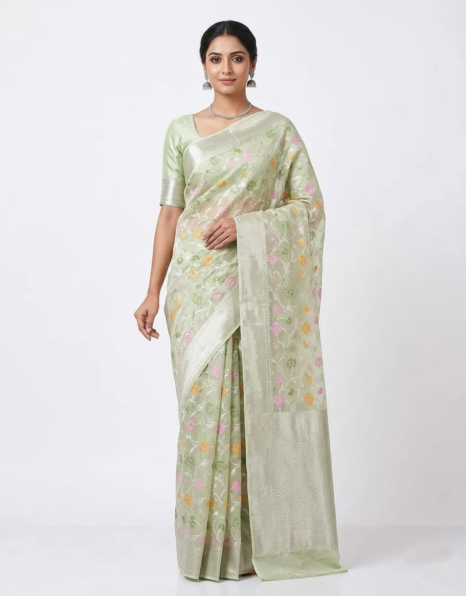 An Indian woman modeling a light sea green organza saree with pink and yellow floral prints and a silver zari border. She is wearing a matching half-sleeved blouse and silver jewelry against a plain white background.
