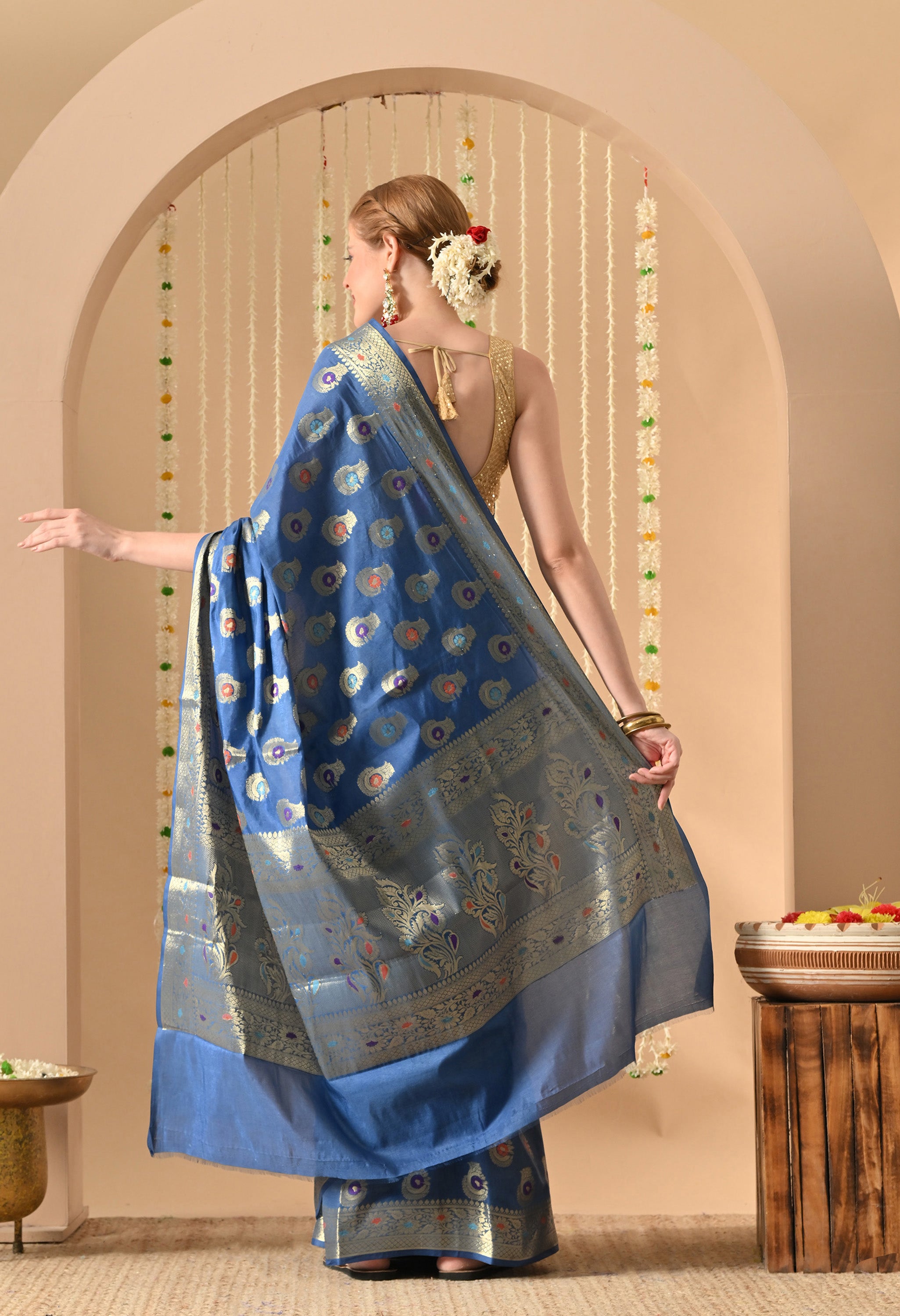 The back view of the The Weavers Shop by Raavi blue silk saree, highlighting the elaborate Zari work on the pallu (decorative end). The model is wearing  gold blouse and her hair styled in a bun. The intricate metallic weaving of the Tilfi patterns is clearly visible across the drape against the minimalist arched backdrop.