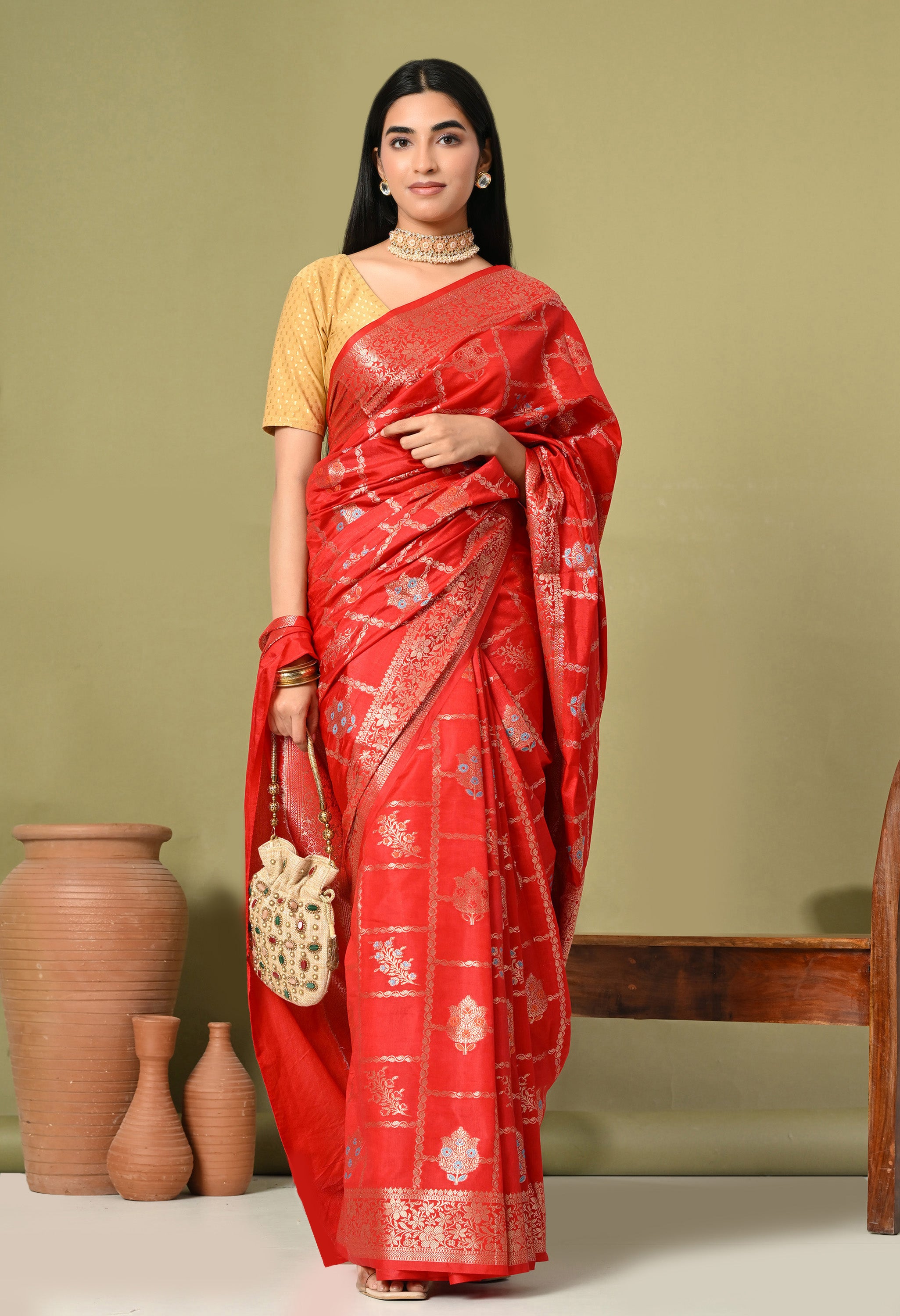 A woman stands against an olive green backdrop, modeling a vibrant red Banarasi silk saree from The Weavers Shop by Raavi. The saree features intricate gold and silver Tilfi work, with a delicate grid pattern framing floral and leaf motifs across the body. It is finished with a wide, ornate gold zari border.

She pairs the saree with a contrasting gold short-sleeved blouse that has a subtle textured pattern. Her accessories include a heavy pearl and gold choker necklace, matching stud earrings, and a tradit