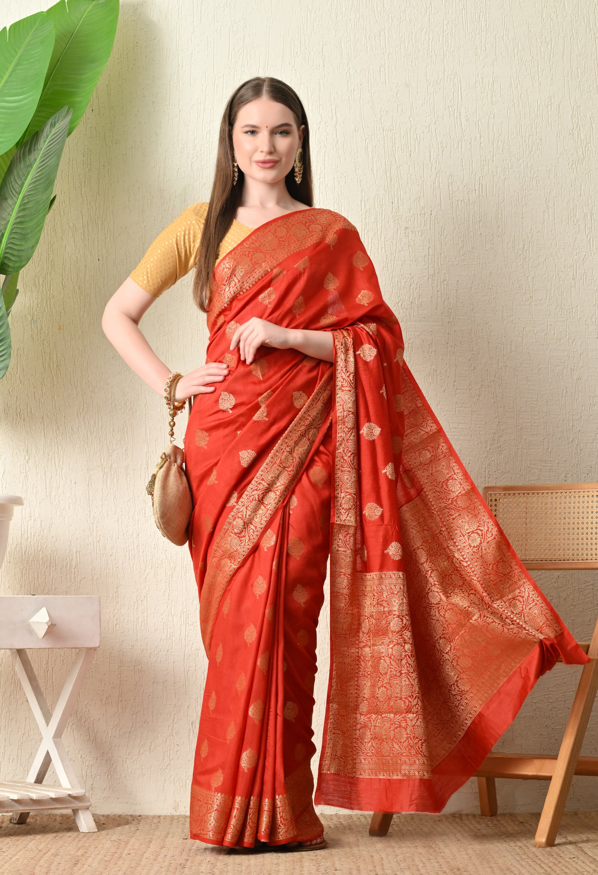 A woman elegantly draped in a luxury red Banarasi silk saree with intricate gold zari work, priced under 3500. The saree features a pattern of small gold leaf motifs across the body and a broad, detailed gold border. She is wearing a short-sleeved gold blouse, holding a matching gold potli bag, and standing in a minimalist interior setting with a large green plant and wooden furniture.