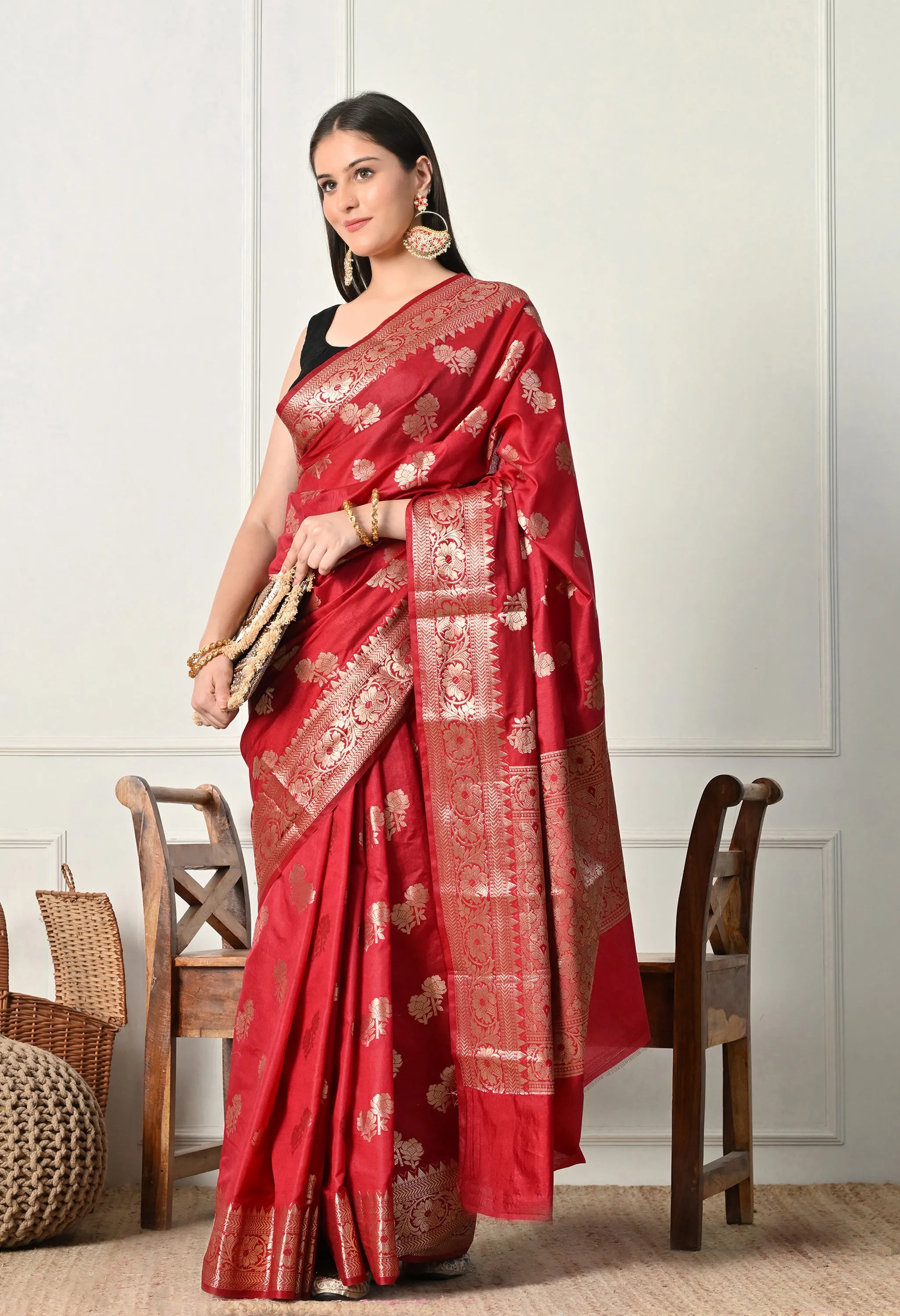 A woman elegantly draped in a crimson red Banarasi silk saree from The Weavers Shop by Raavi. The saree features intricate golden zari work, including traditional floral buttas across the body and a heavy, ornate gold border and pallu. She is styled with a sleeveless black blouse, statement earrings, and a matching clutch, standing against a neutral white wall with rustic wooden furniture.