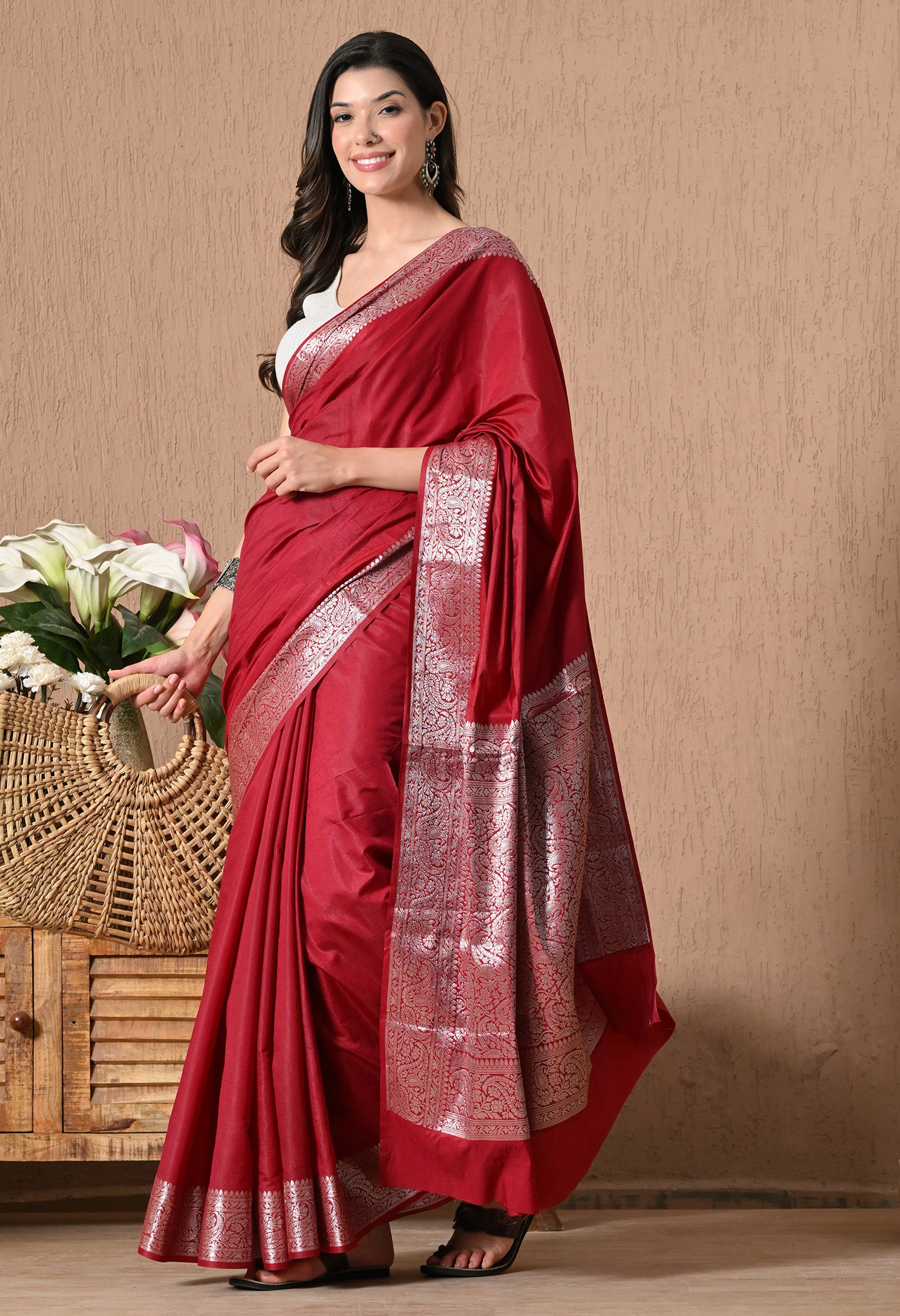A full-length front view of a woman modeling a deep red silk saree by The Weavers Shop by Raavi. The saree features a minimalist solid body contrasted by a thick, ornate silver zari border and pallu. She is styled with a white crop-top blouse and statement silver earrings, standing in a warm, decorated indoor setting.