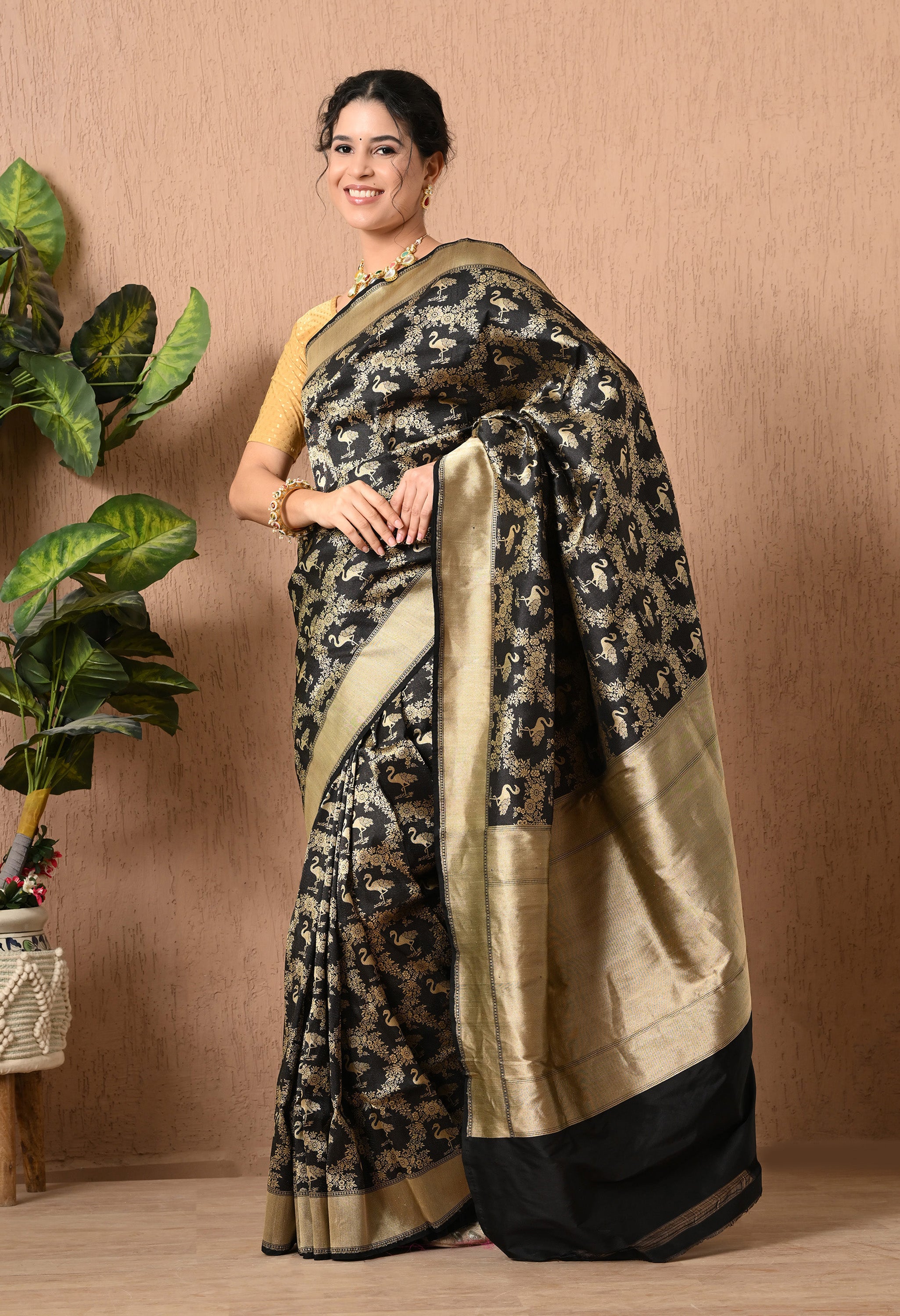 A full-length front view of a smiling woman posing in an elegant black handwoven zari Shikarga saree from The Weavers Shop by Raavi. The Katan Silk fabric is densely covered in a shimmering gold pattern featuring traditional Shikarga motifs of animals and birds. This Silk Mark Certified saree is paired with a mustard-gold textured blouse, a heavy stone-encrusted necklace, and matching bangles, creating a luxurious look for weddings or receptions.