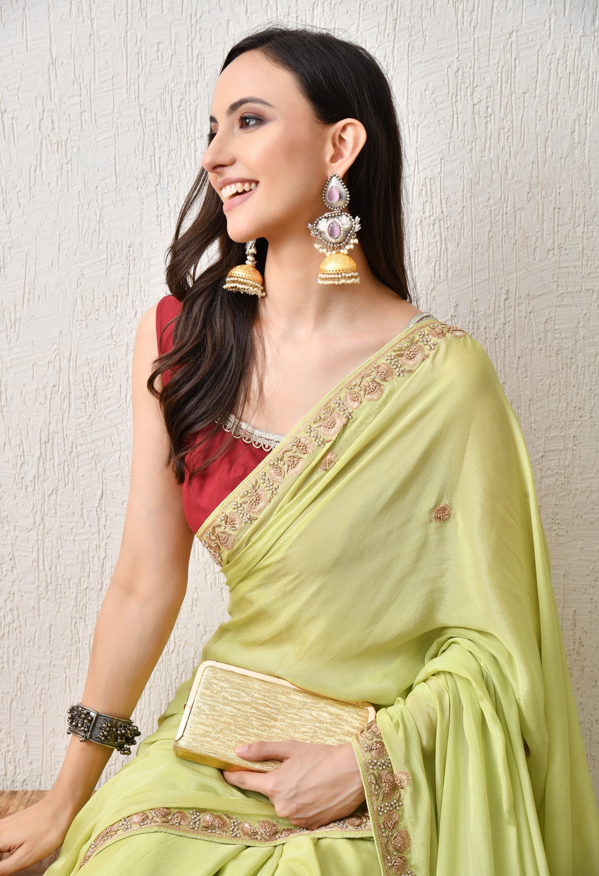 A side-profile close-up of a woman, wearing a pista green chiffon saree from The Weavers Shop by Raavi. The shot emphasises her large, elaborate gold and silver jhumka earrings and the delicate Zardosi border of the saree. She is holding a gold clutch bag.