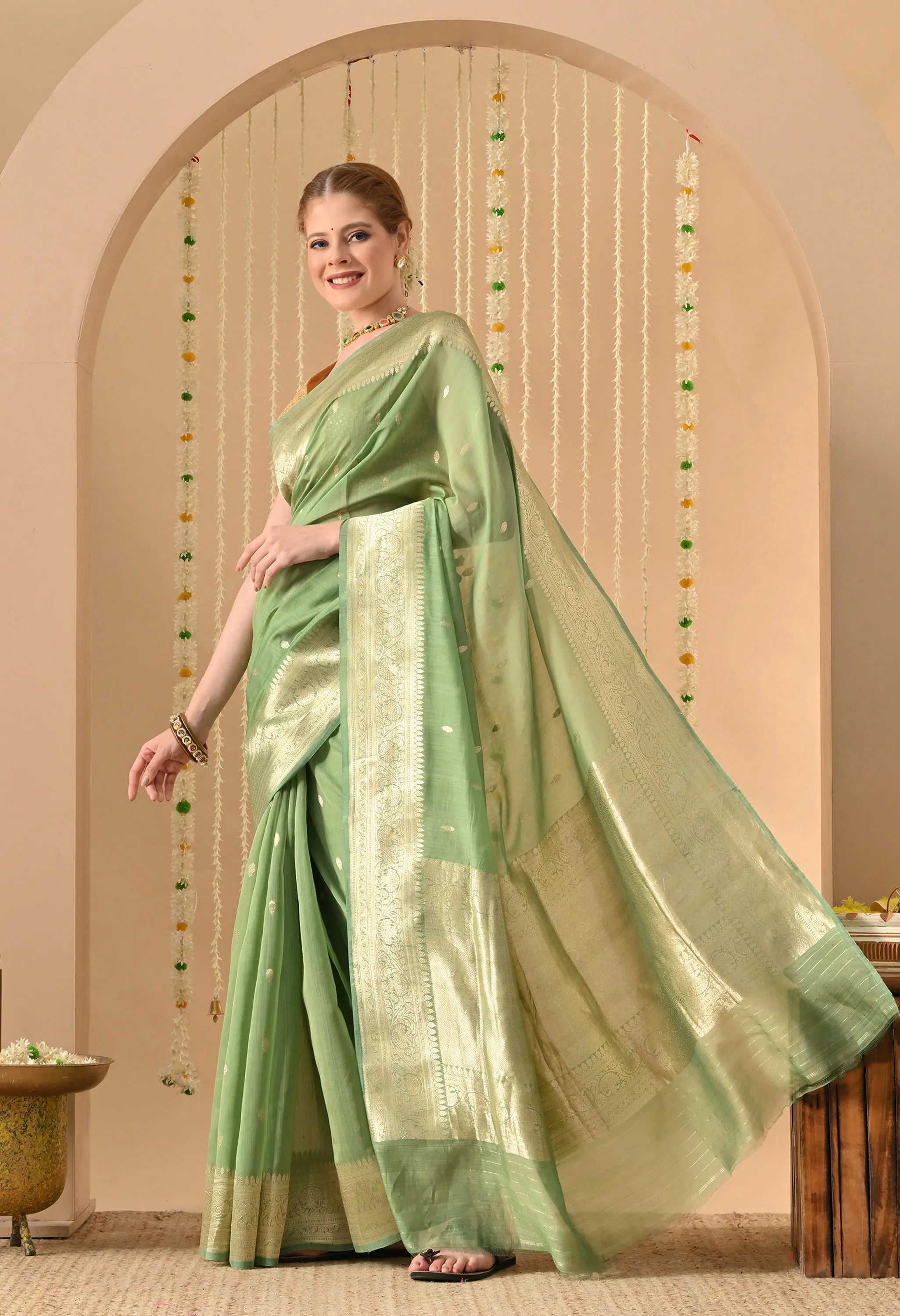 A side-profile shot of a model showcasing the luxurious drape of a Silk Mark Certified Mint Green Pure Silk Saree from The Weavers Shop by Raavi. The image highlights the hand-woven Karuwa Weave, featuring small gold zari work motifs that stand out against the rich, opaque texture of the pastel silk. The wide, shimmering gold border is displayed in full detail as it flows elegantly from the shoulder, emphasizing the artisanal craftsmanship and sophisticated silhouette of this Banarasi masterpiece.