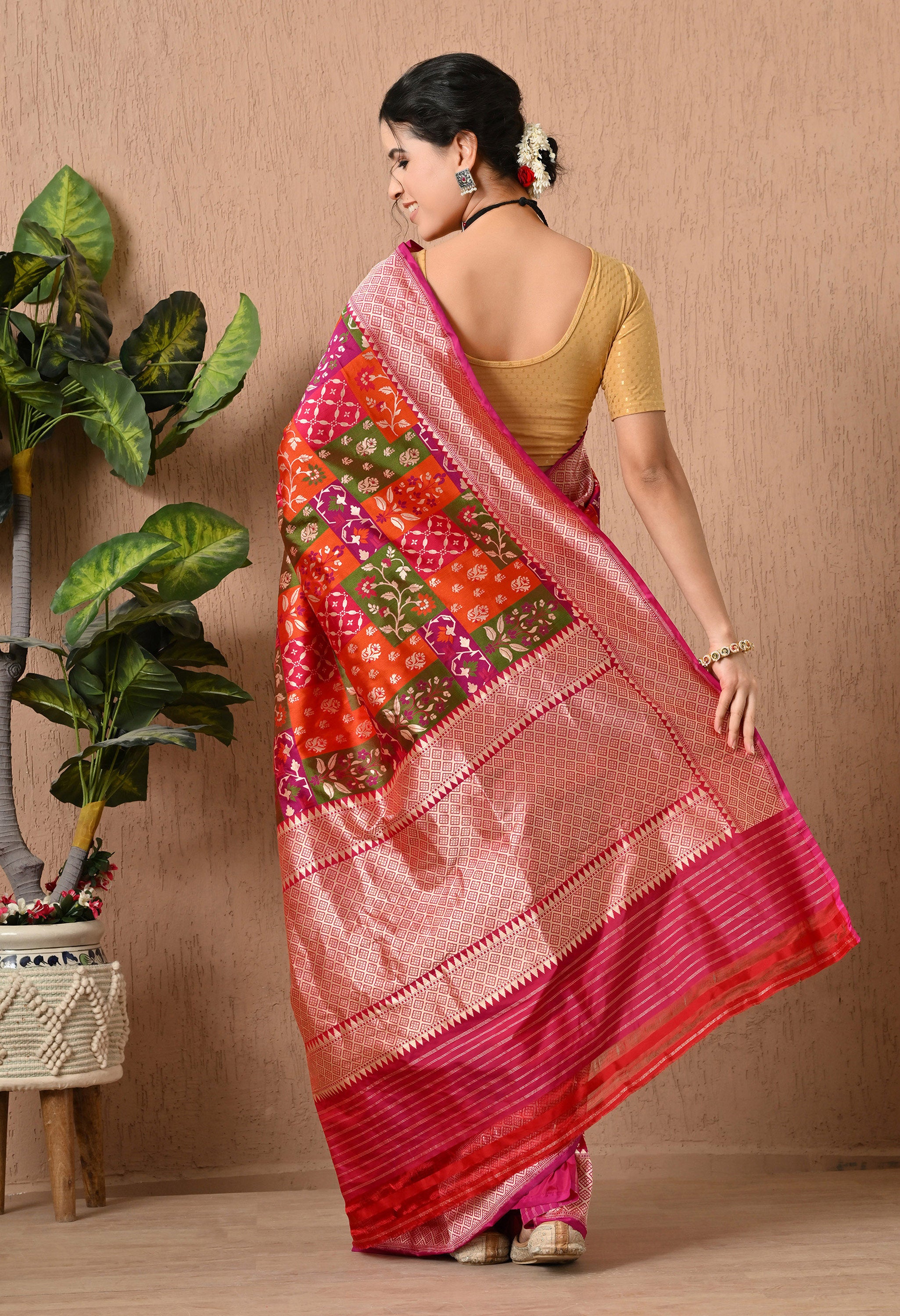 A back-angle view of a woman showcasing the elaborate pallu of a multi-colored Rangkat saree by The Weavers Shop by Raavi. The image highlights the dense gold zari work woven into a geometric diamond grid across the pallu, finished with horizontal zari stripes at the edge. The Pure Katan Silk fabric displays a beautiful sheen, contrasting the bright orange, green, and magenta handwoven panels. The woman has her hair in a bun adorned with white flowers (gajra) and is wearing a gold blouse and statement earri