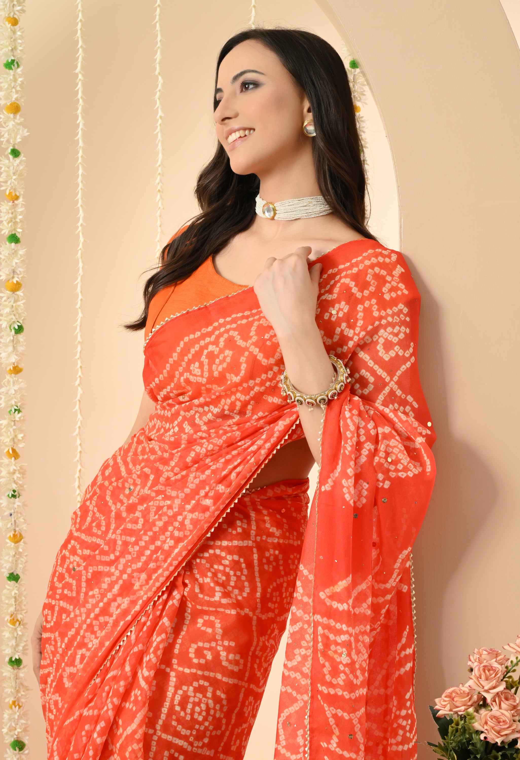 A side-angle close-up of a smiling woman wearing a vibrant orange georgette saree from The Weavers Shop by Raavi. This handcrafted Bandhani saree, perfect for a festive event or a Bollywood-themed party, features a traditional white tie-dye pattern and a delicate gold-trimmed border. She is accessorized with a pearl choker, matching earrings, and a statement bangle, leaning against a neutral-toned wall decorated with jasmine garlands.