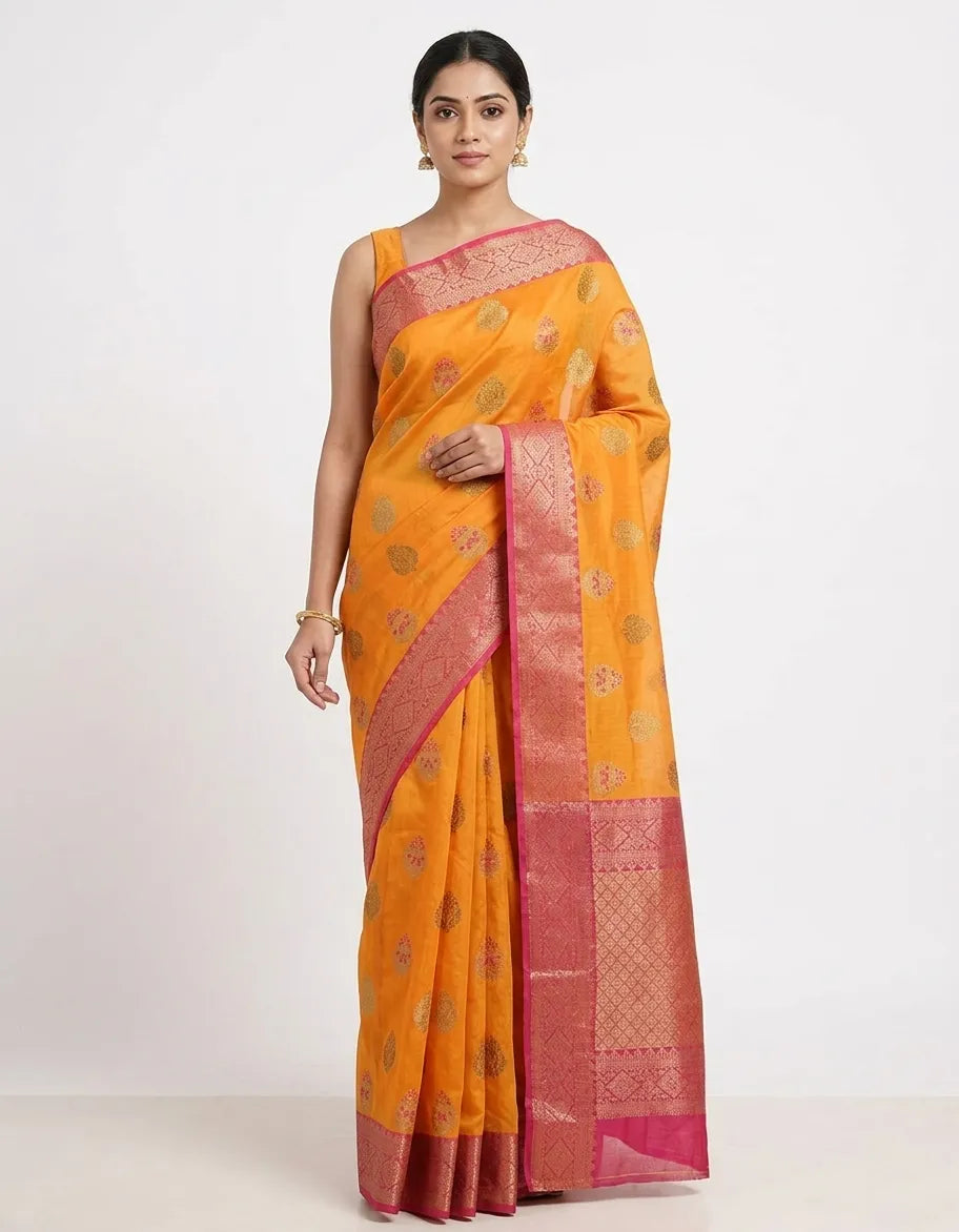 An Indian woman modeling a mustard yellow silk blend saree with gold woven motifs and a contrasting deep pink zari border. She is wearing a sleeveless yellow blouse and gold earrings against a plain white background.

