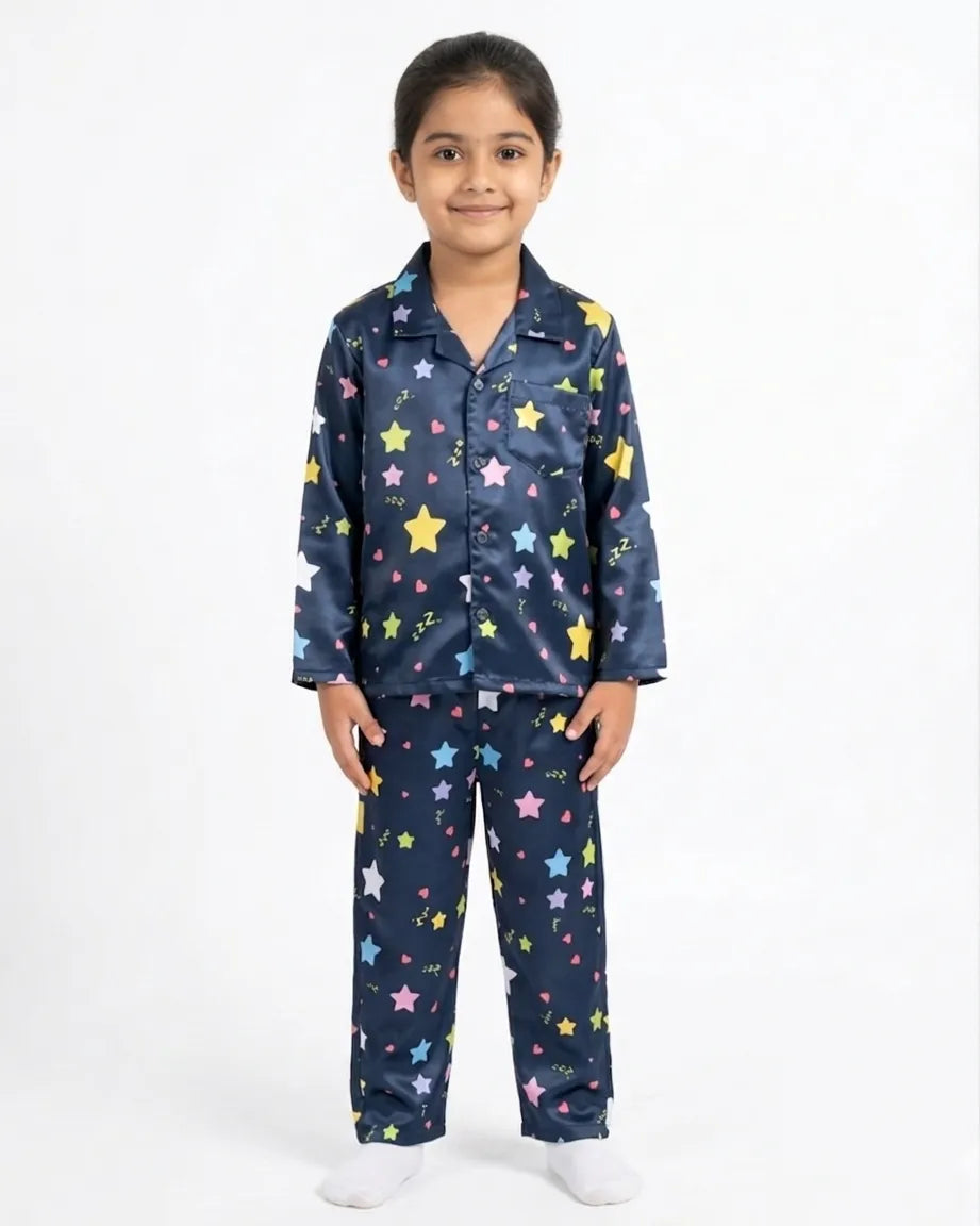 A young girl smiling against a plain white background, wearing a navy blue satin night suit with a vibrant, colorful all-over star and heart print. The two-piece pajama set features a button-down shirt with a collar and matching full-length trousers.