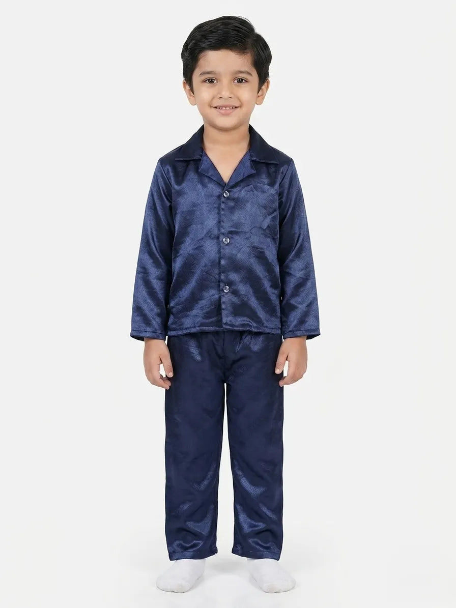 A young boy smiling and wearing a shiny navy blue satin night suit set featuring a long-sleeved buttoned shirt and matching full-length pyjamas against a white background.
