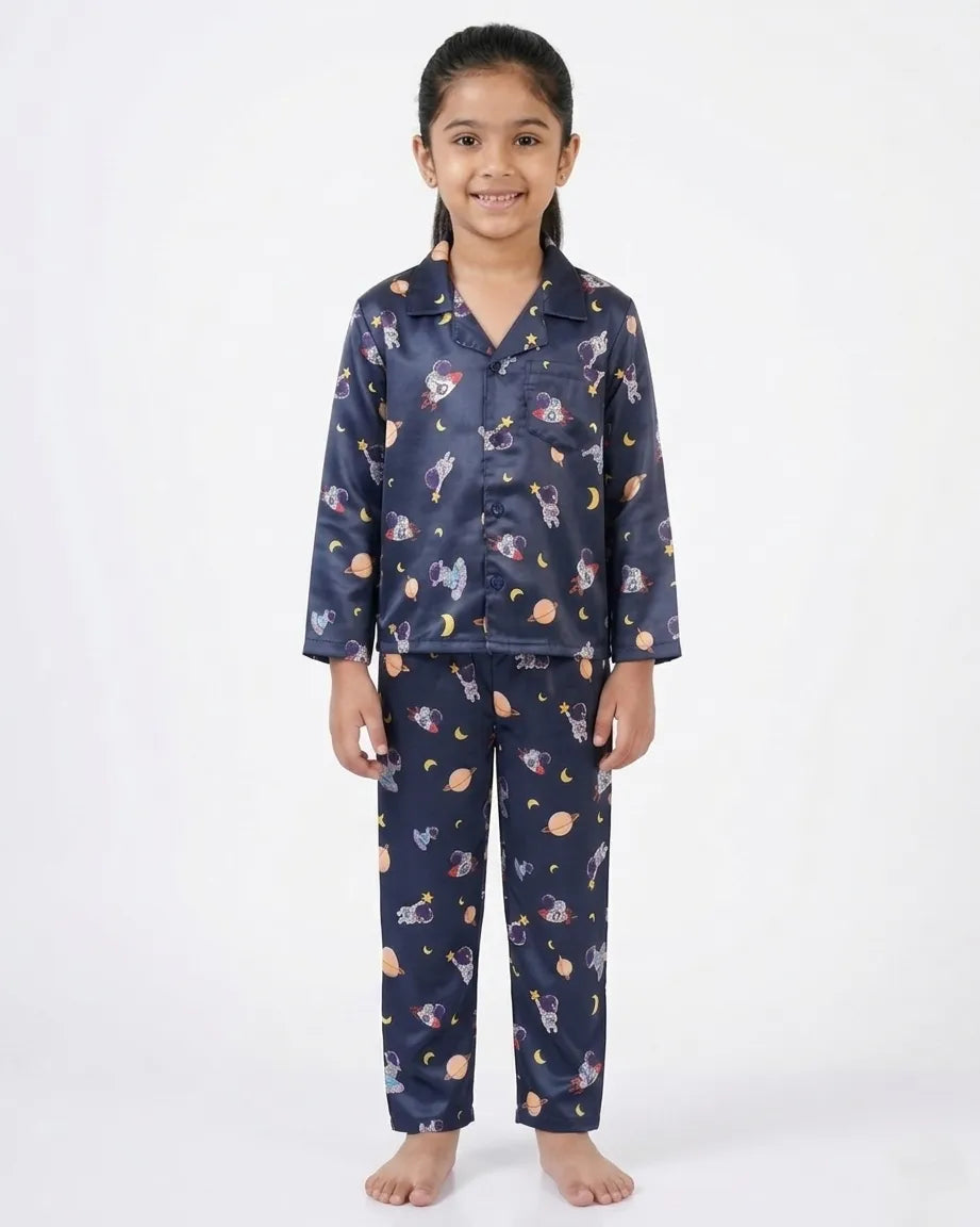 A young girl smiling against a white background, wearing a navy blue satin night suit with a playful all-over space-themed print featuring planets, stars, and rockets. The set includes a long-sleeved button-down shirt with a collar and matching full-length pajama trousers.