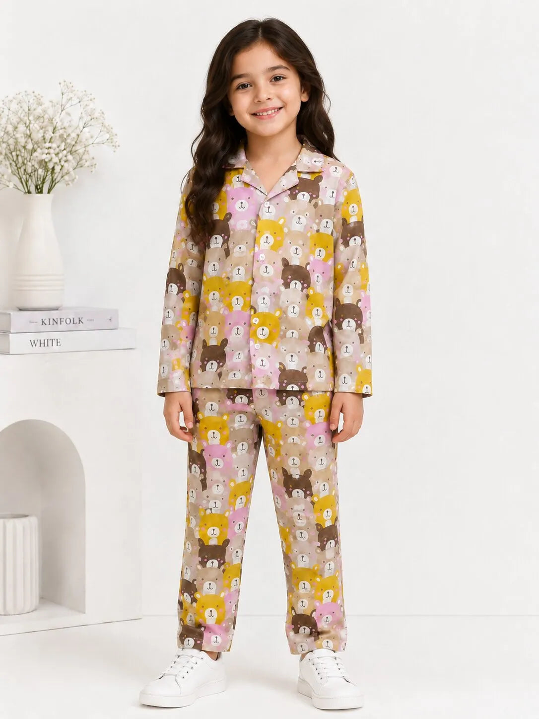 A young girl smiling while standing against a bright, minimalist white background, wearing a matching two-piece pajama set. The long-sleeve button-down shirt and trousers feature a colorful, repeating cartoon bear pattern in shades of brown, yellow, and pink on a beige base. She is paired with clean white sneaker