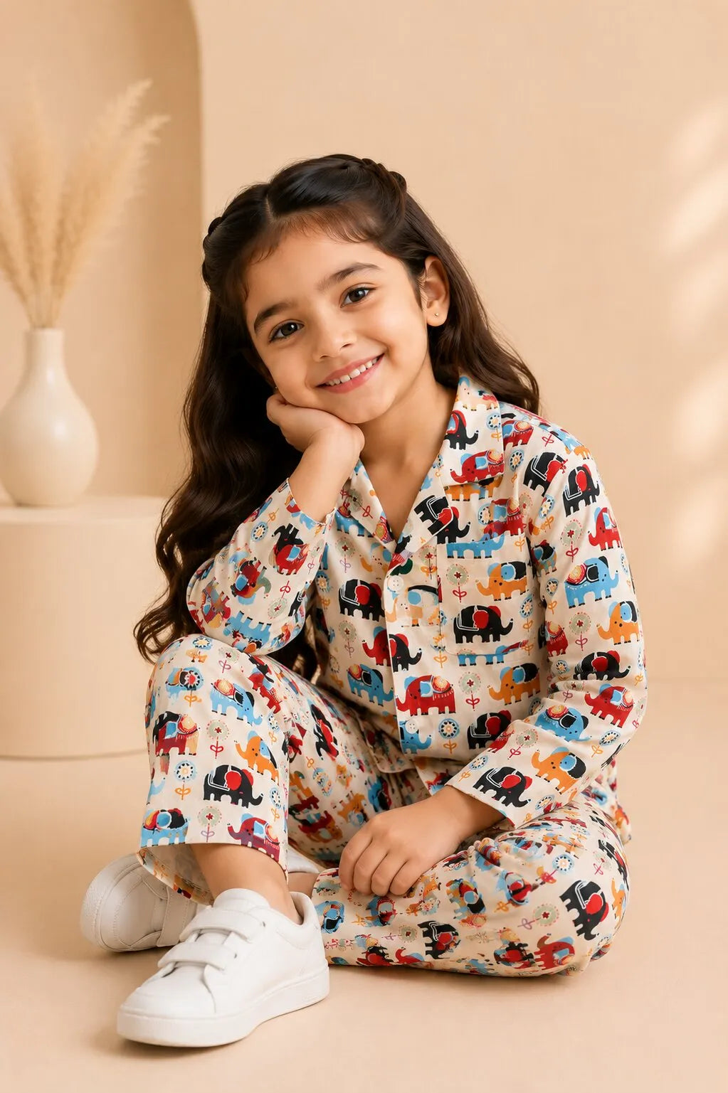 A young girl  smiling in a cozy bedroom, wearing a beige cotton night suit with a colorful red and blue elephant print.
