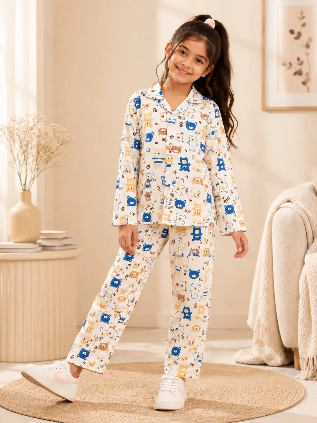 A young girl sitting cross-legged wearing a white satin night suit with a blue and brown bear print.
