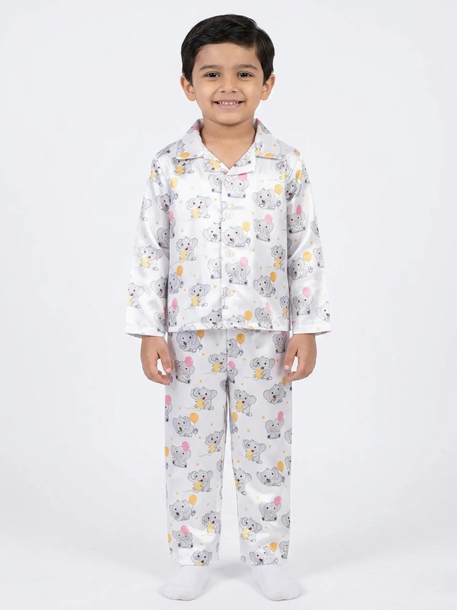 A smiling young boy wearing a white satin night suit featuring a cute grey elephant and colorful balloon print.
