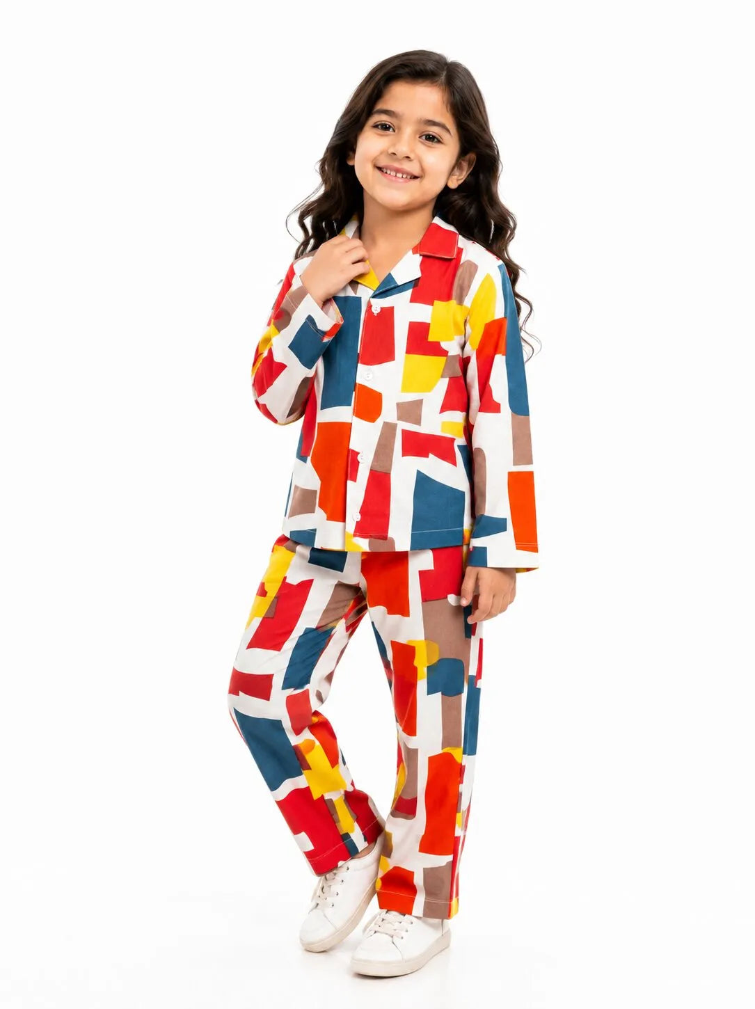 A young girl standing against a white background, wearing a white long-sleeved button-up pajama set with a colorful, multi-colored geometric square print