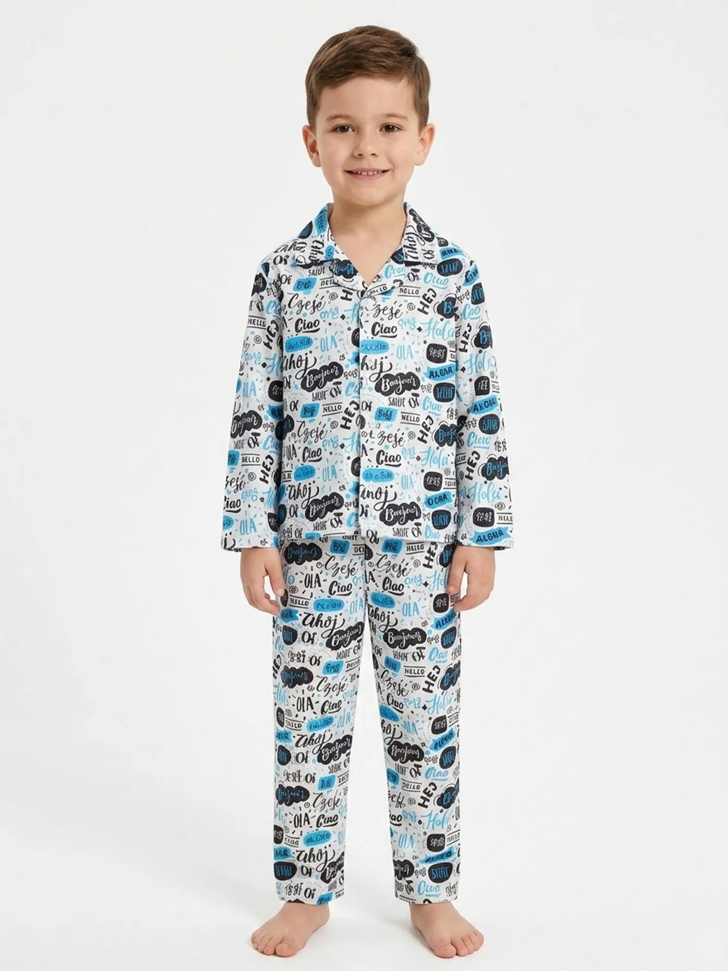 A young boy standing against a white background wearing a white satin night suit with a blue and black multilingual "Hello" typography print.
