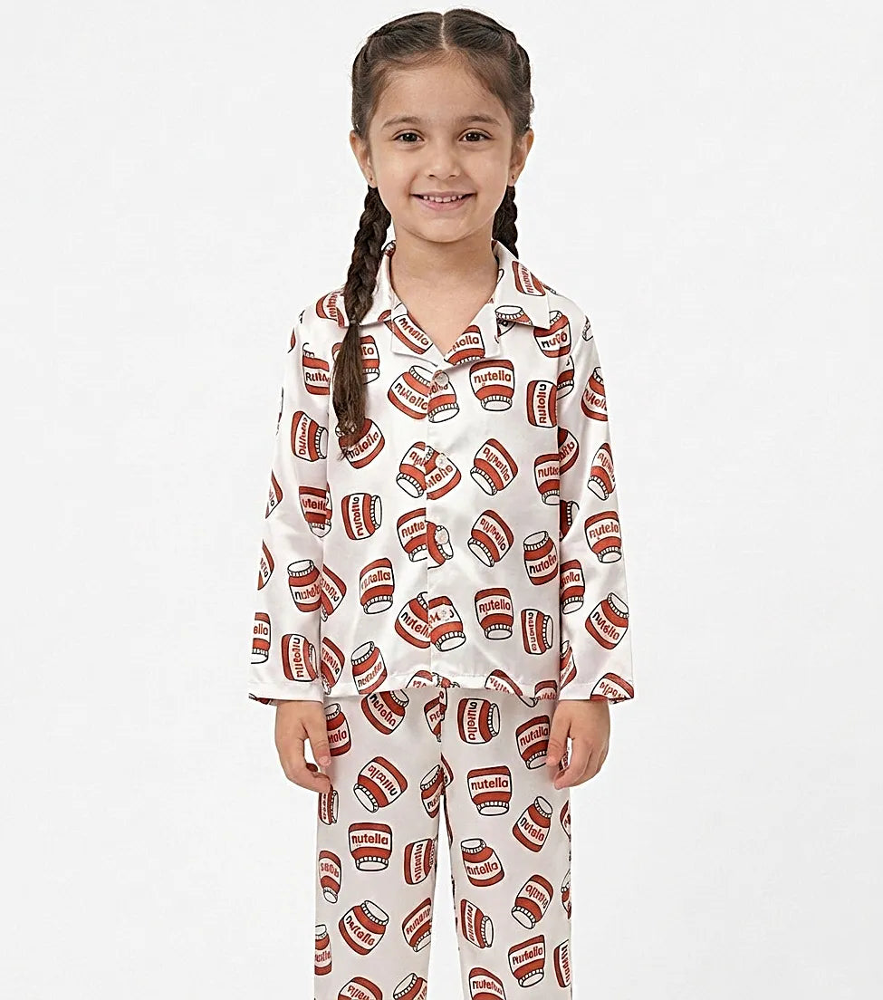 A young girl standing against a white background wearing a white satin long-sleeved night suit with a red hazelnut spread jar print.
