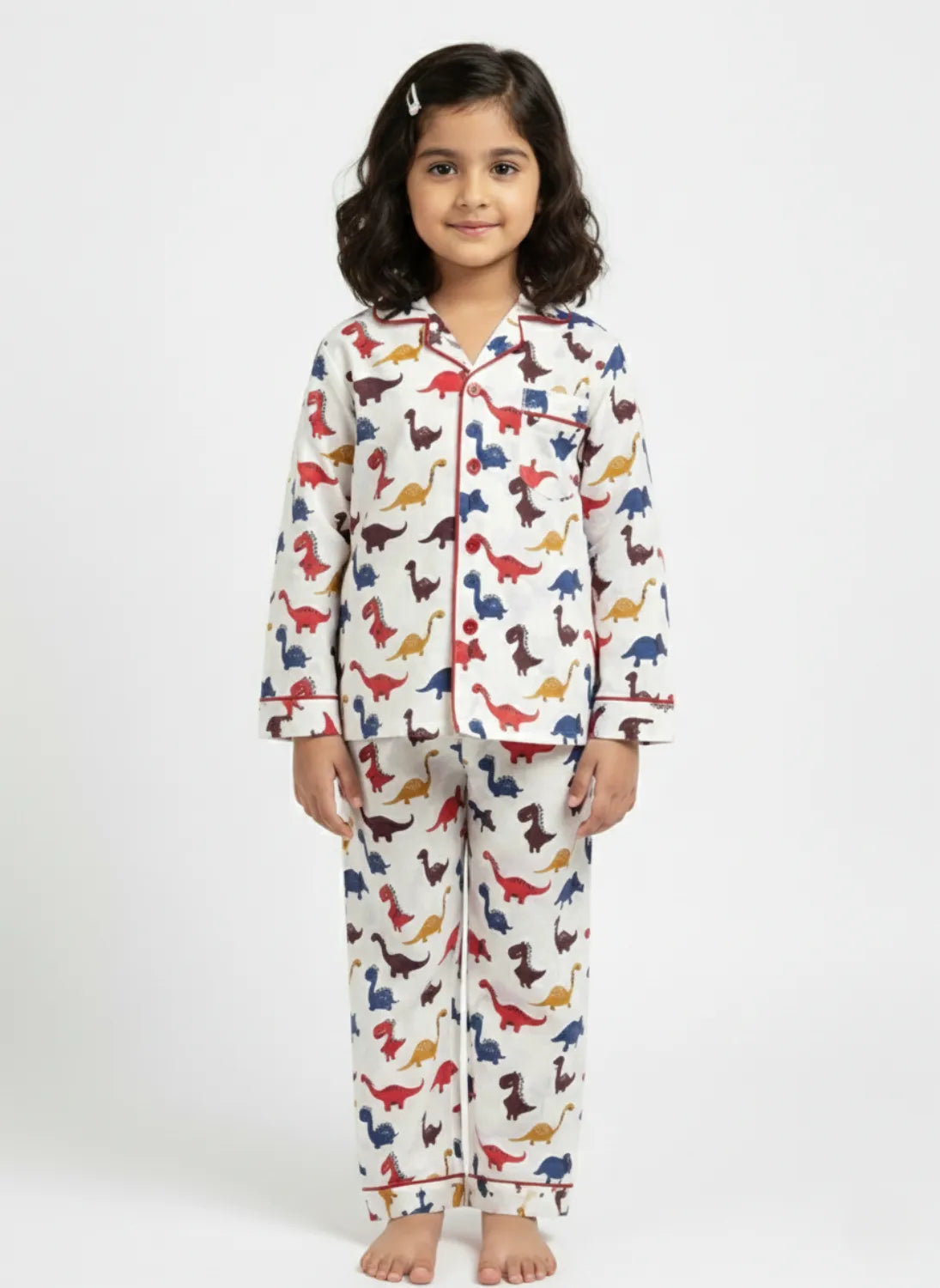 A young girl standing against a white background, wearing a white long-sleeved button-up pajama set with a colorful all-over dinosaur print