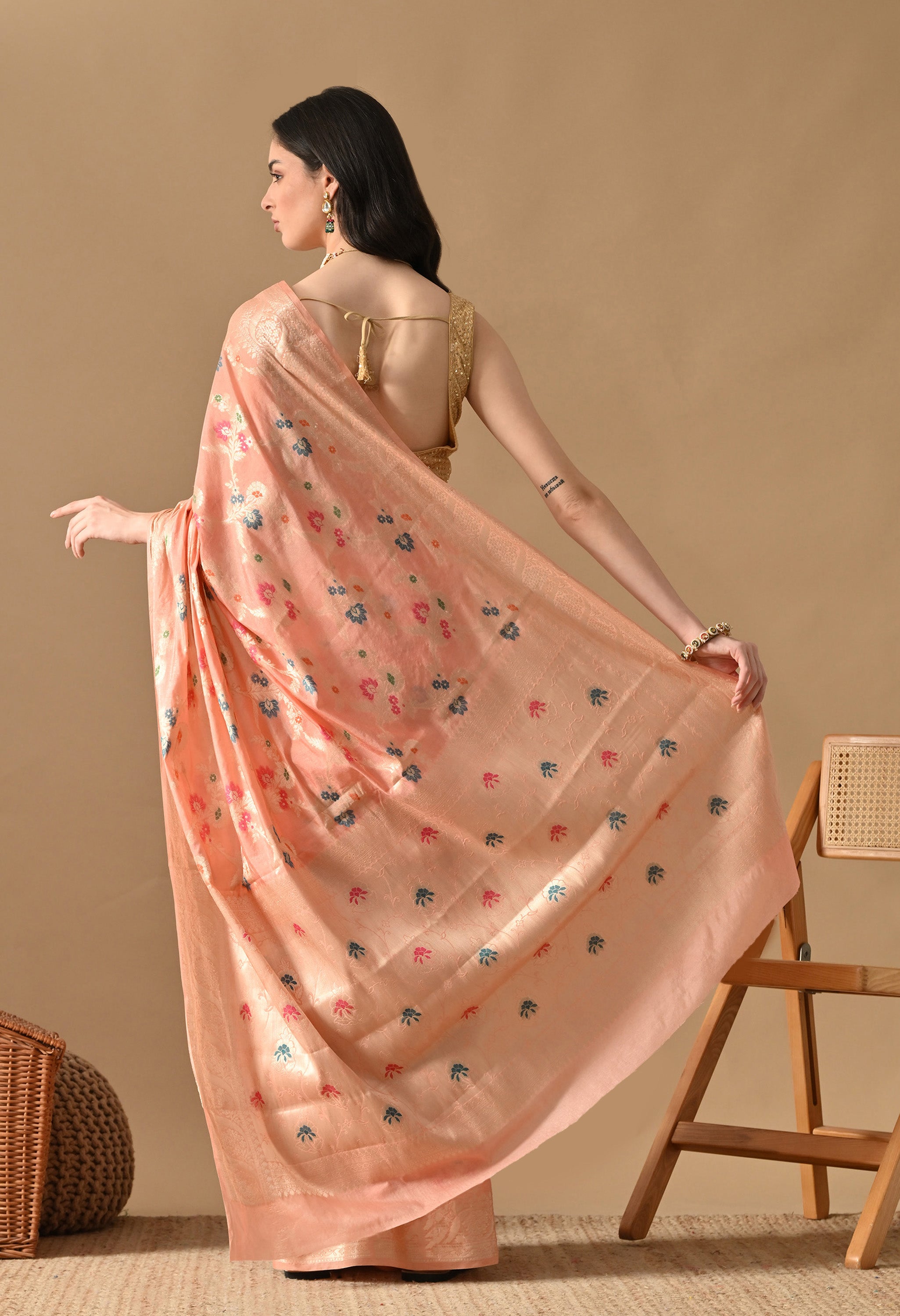 A woman is shown from the back and side, highlighting the drape of a peach silk saree by The Weavers Shop by Raavi. The Zari and Tilfi work floral patterns are visible across the back of the saree, which is paired with a gold sleeveless blouse featuring tie-back detailing. The image showcases the shimmering gold zari border as she holds the edge of the machine-woven fabric.