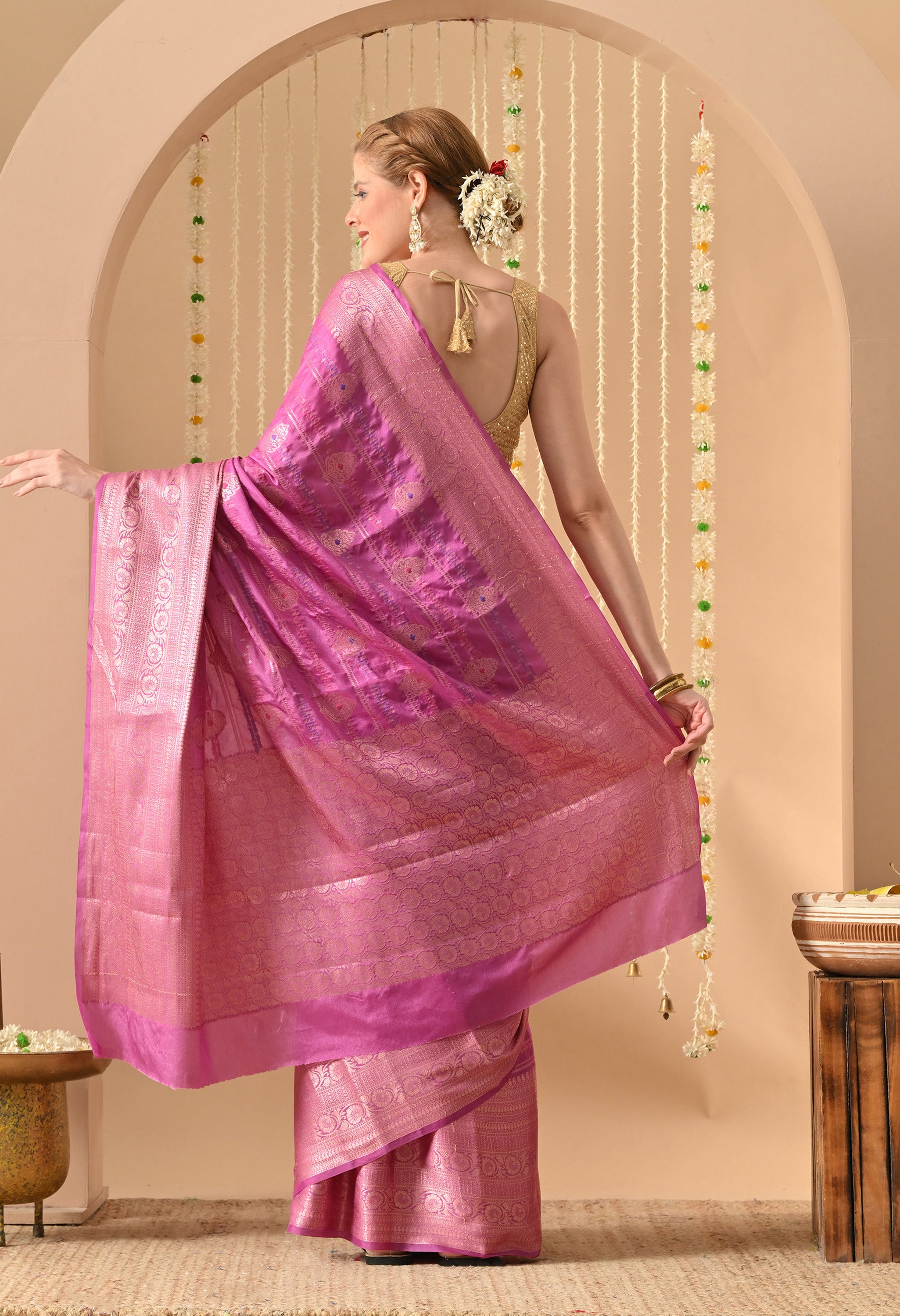 A back view of a model showcasing the exquisite craftsmanship of The Weavers Shop by Raavi. She is draped in a vibrant magenta Banarasi silk saree, highlighting the extensive Tilfi work and Zari detailing on the pallu and body. The saree is paired with a gold sequined backless blouse featuring tassel ties, set against a festive backdrop of white flower hangings and golden bells.