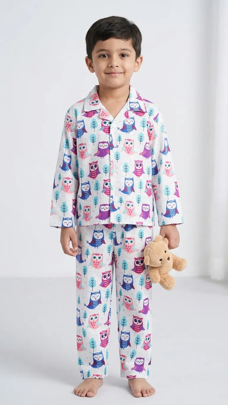 A young boy smiling and holding a teddy bear, wearing a white two-piece pajama set with a colorful, all-over owl print in shades of pink, purple, and blue.