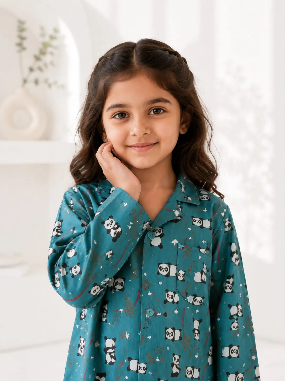 A young girl standing on a light wooden floor, wearing a teal blue satin night suit with a playful panda and bamboo print. The two-piece pajama set features a long-sleeved button-down shirt with a notched collar and matching full-length trousers.