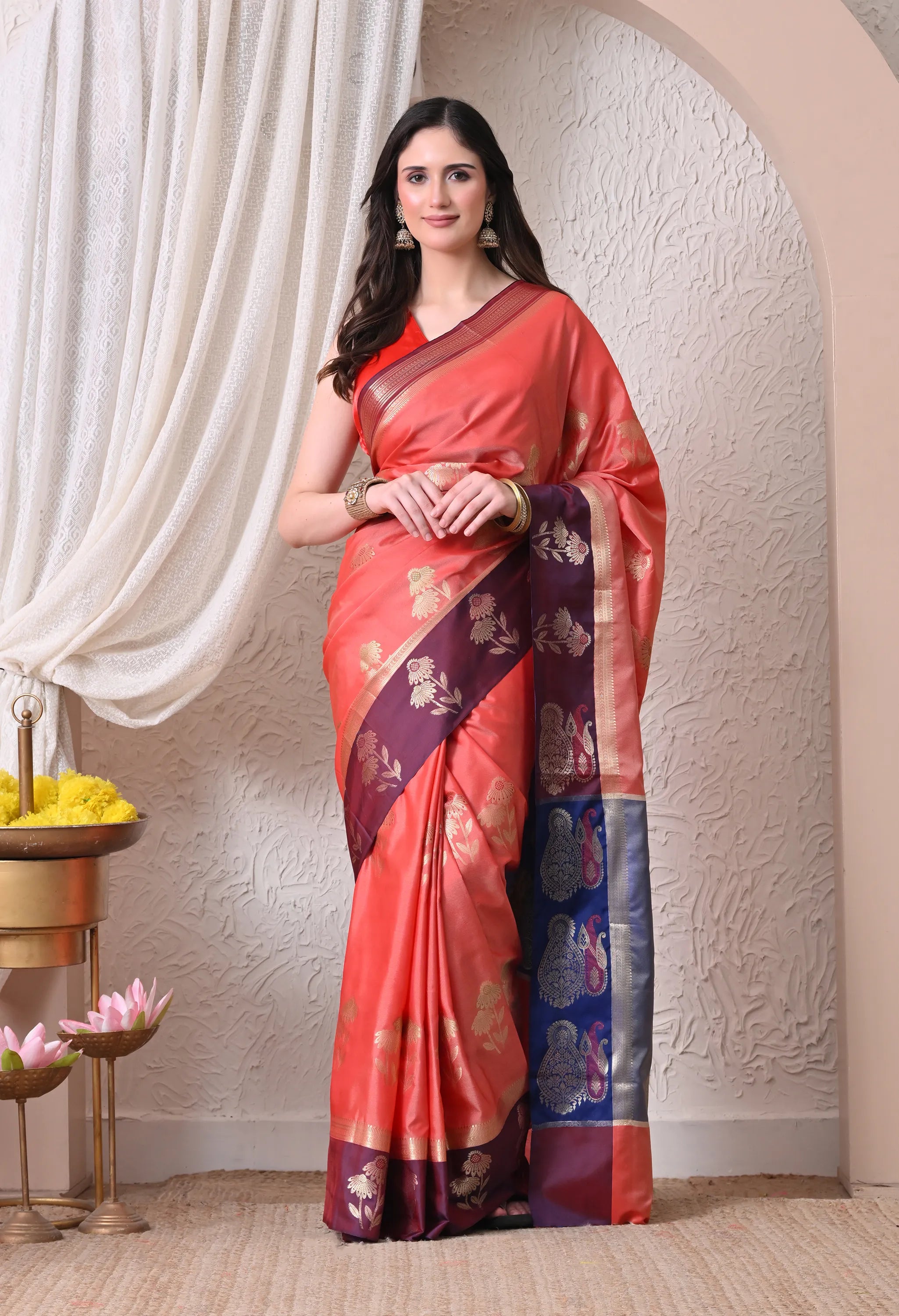 A woman posing in a vibrant peach-coral silk blend saree featuring golden floral motifs and a contrasting deep wine and royal blue woven border and pallu.
