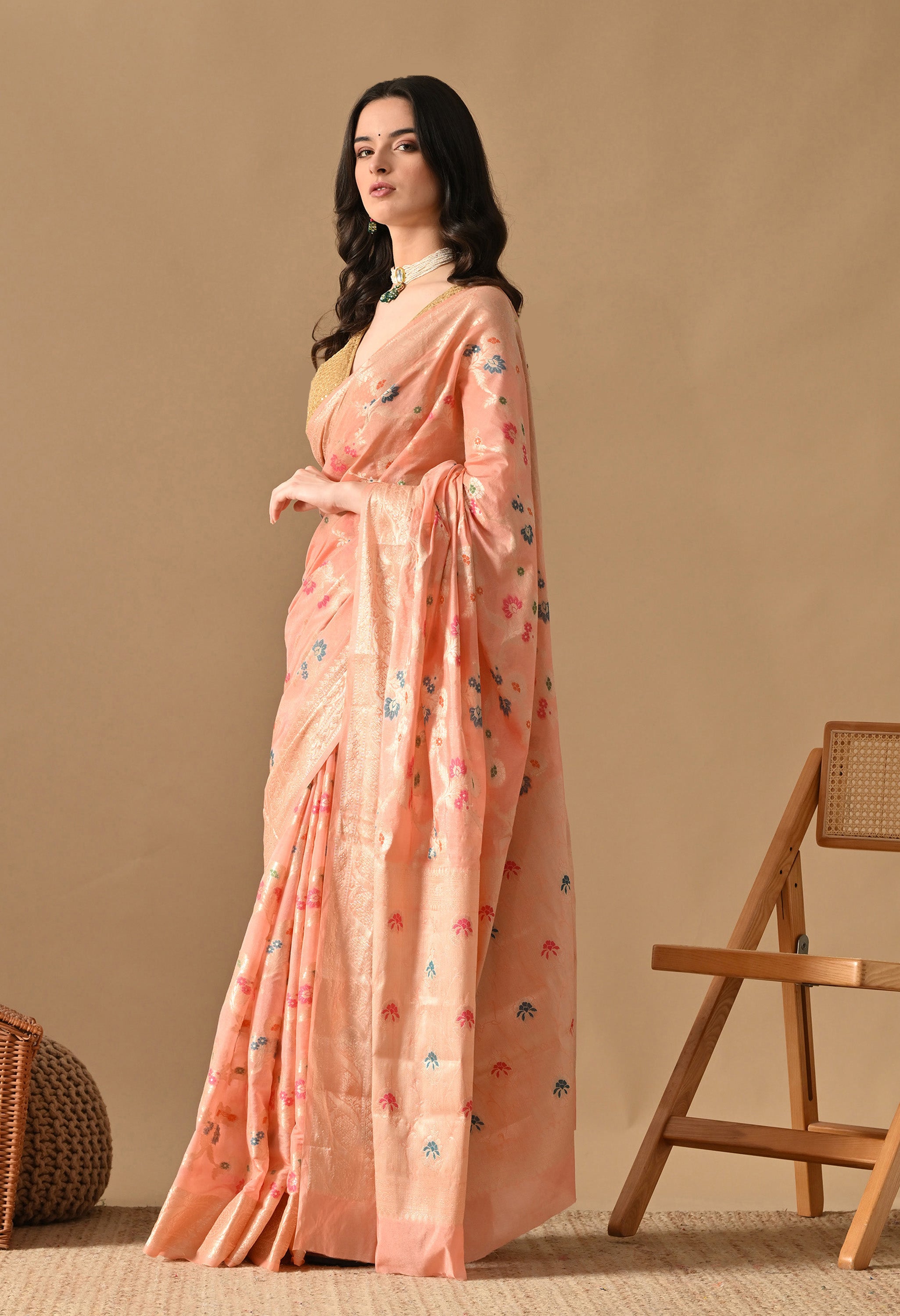 A woman is captured in a three-quarter pose wearing a peach silk saree from The Weavers Shop by Raavi. The design features Tilfi work with Zari accents and colourful floral woven work. The saree is paired with a gold sleeveless blouse and a pearl choker. The image showcases the soft drape of the woven, silk fabric against a neutral studio background.