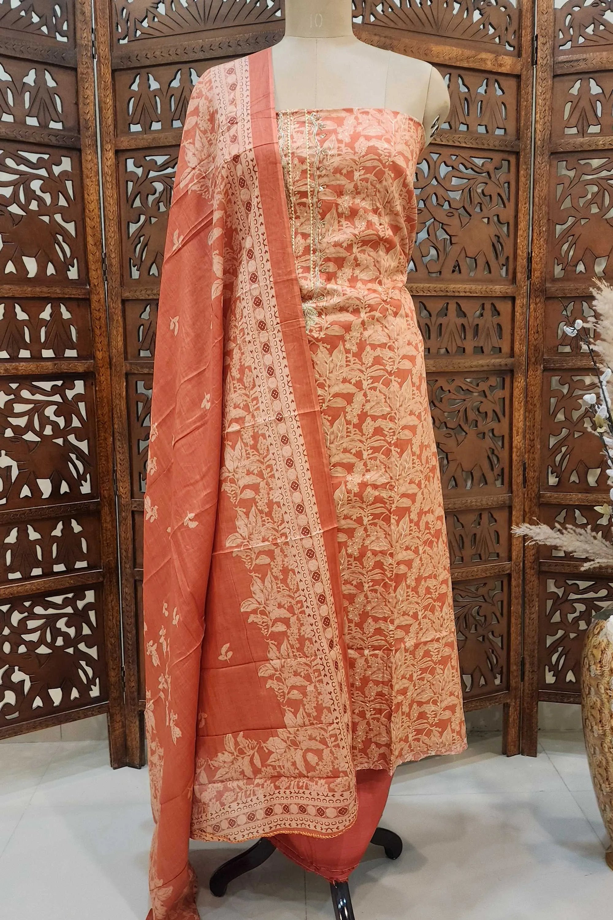 A peach and off-white floral printed unstitched pure cotton suit displayed on a mannequin. The set includes a floral kurta, a matching dupatta with an elegant printed border, and solid peach bottoms, set against a traditional wooden background.