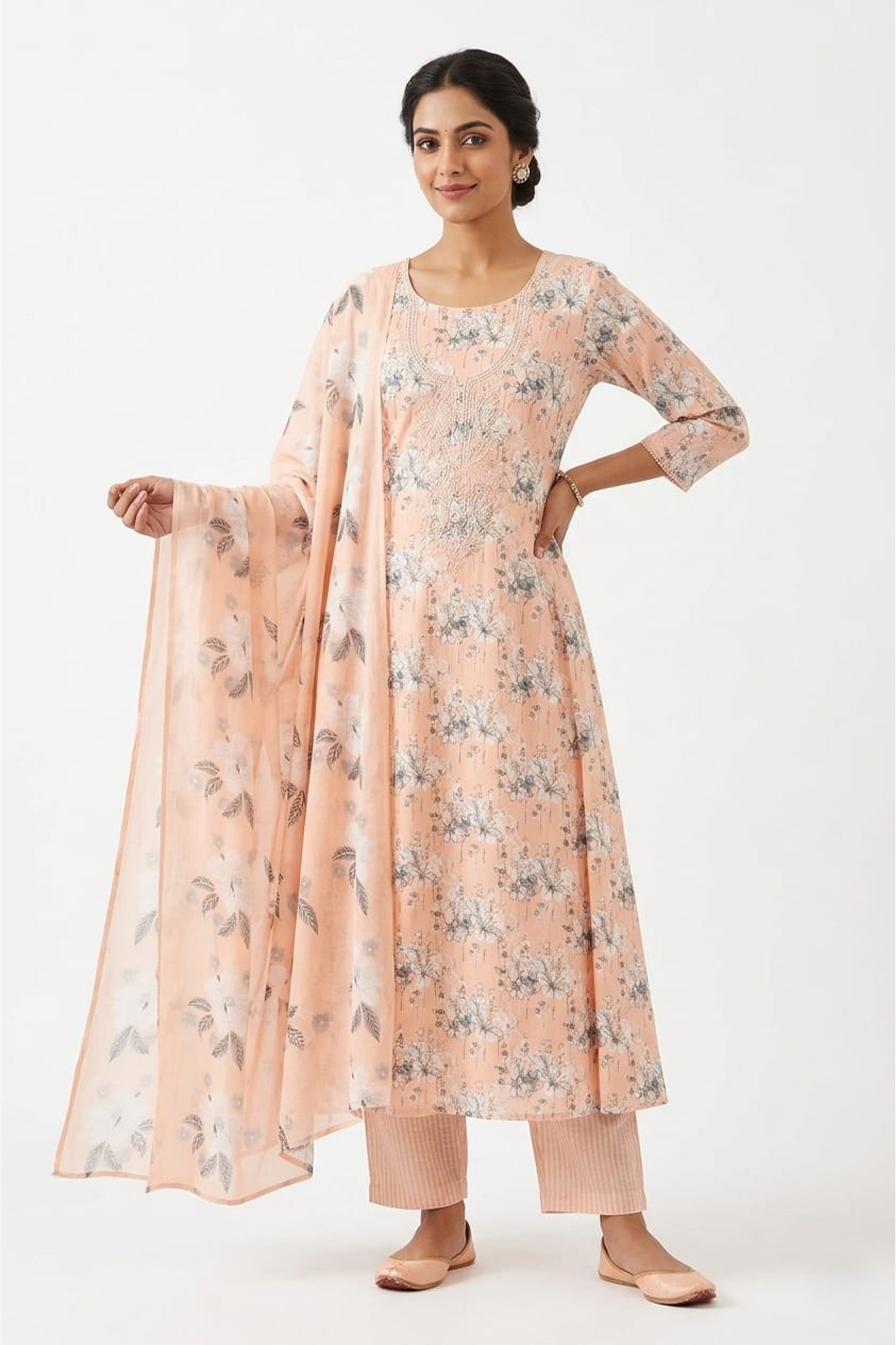 A pastel peach cotton unstitched suit with white floral prints and an embroidered neckline, shown with a matching dupatta and striped bottom fabric.
