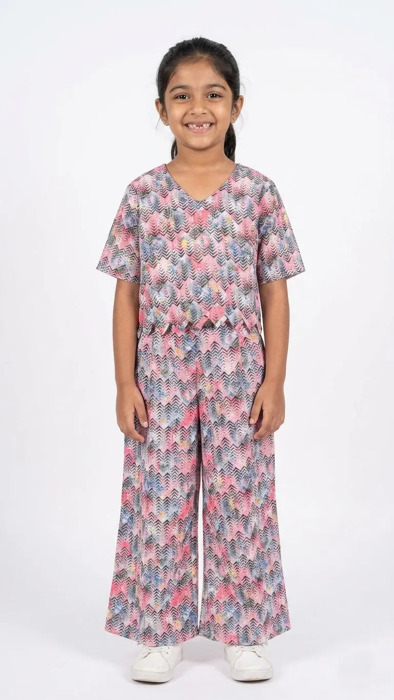A young girl is smiling and posing in a two-piece coordinated set against a plain white background. The outfit consists of a short-sleeved, V-neck top with a scalloped hem and matching wide-leg trousers.