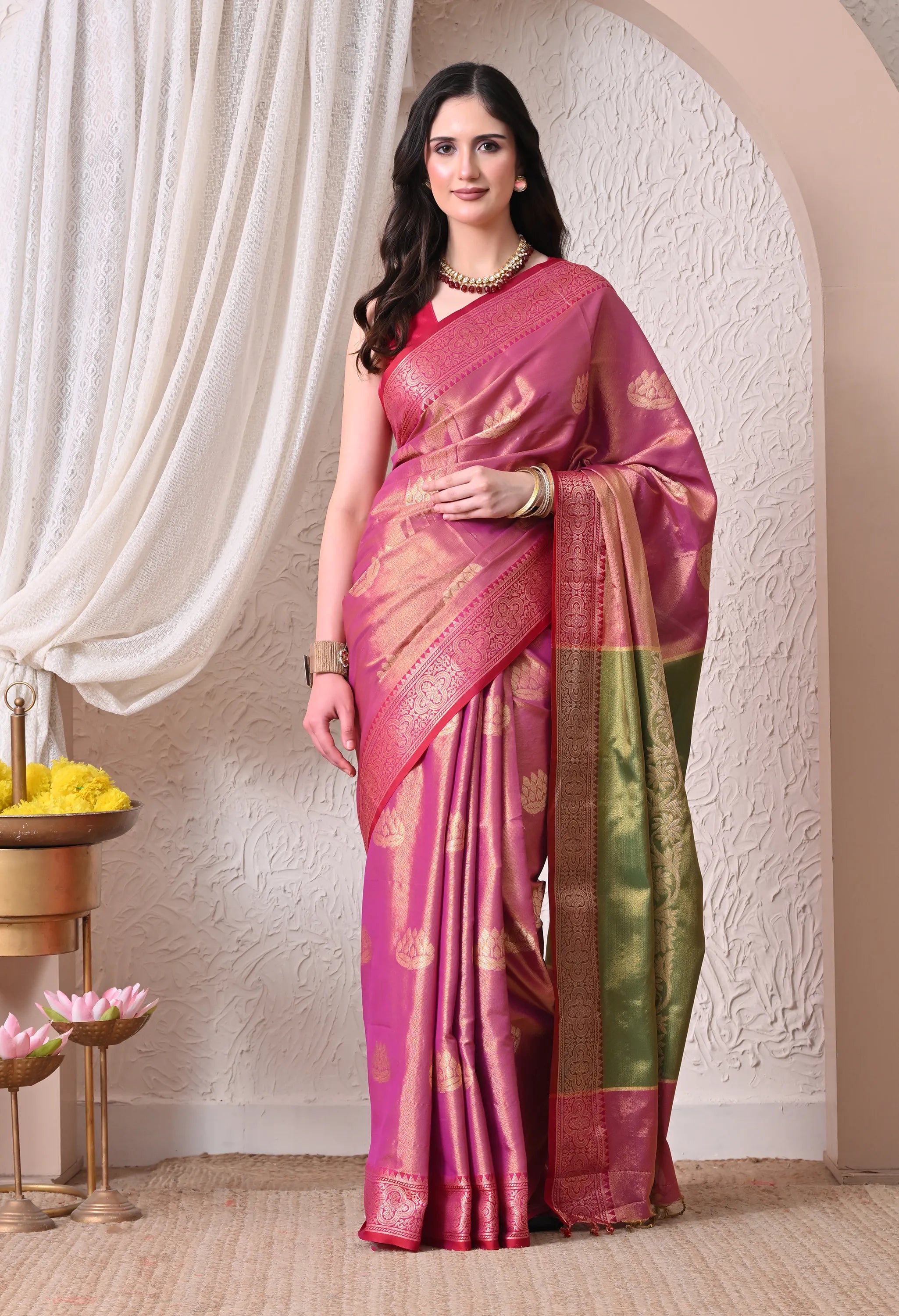 A woman posing in a vibrant Rani Pink silk blend saree featuring woven gold lotus motifs, an intricate olive green and maroon zari border, and a matching gold necklace.
