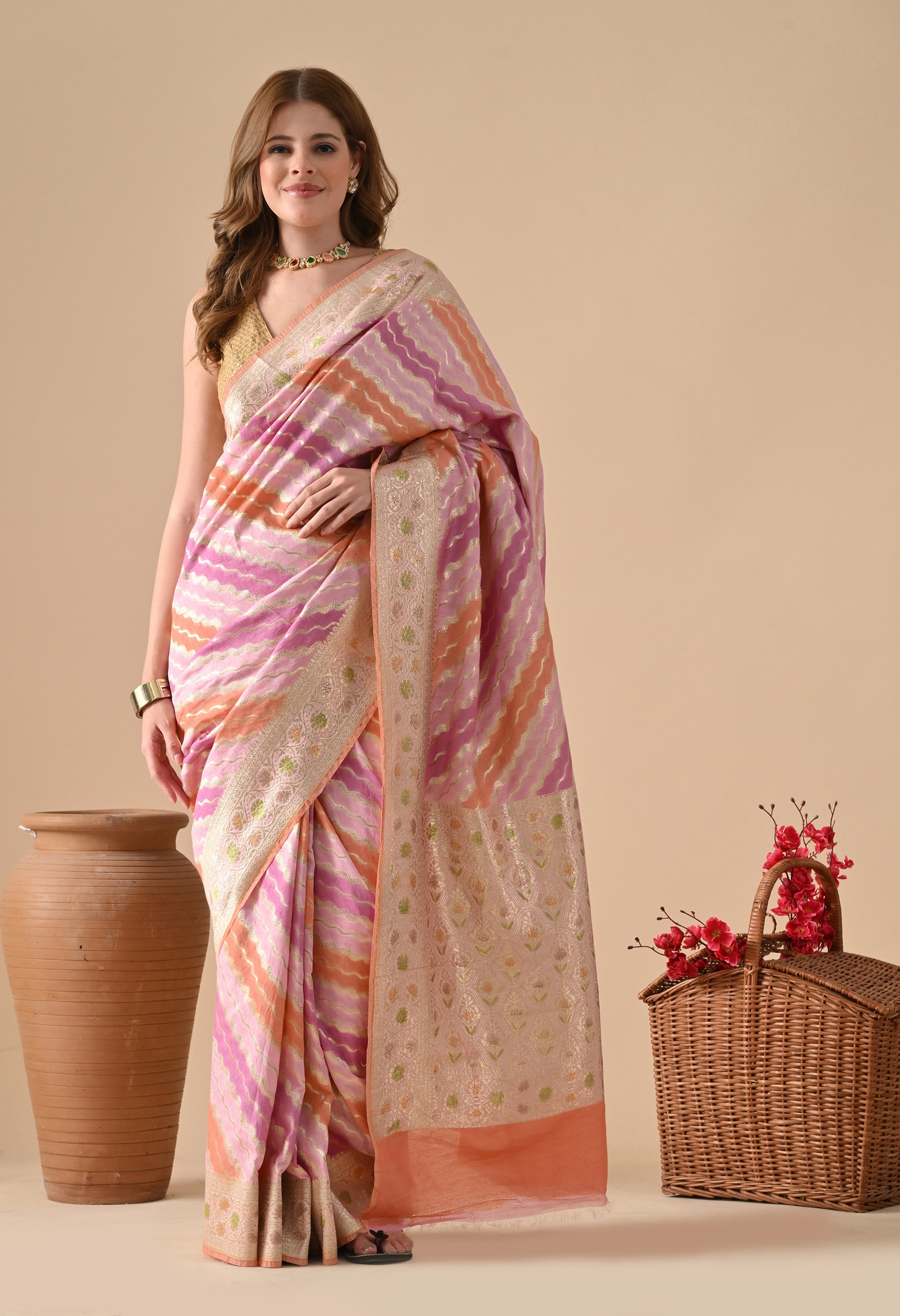 A woman elegantly poses in a The Weavers Shop by Raavi Banarasi silk saree, featuring the signature Rangkat technique. The saree showcases horizontal wavy stripes in pastel pink, lavender, and vibrant orange, woven with shimmering gold zari work. The broad pallu and borders are heavily embellished with intricate gold floral patterns. She is styled with a sleeveless gold blouse and a necklace, standing between a large earthenware pot and a flower-filled wicker basket against a warm neutral backdrop.