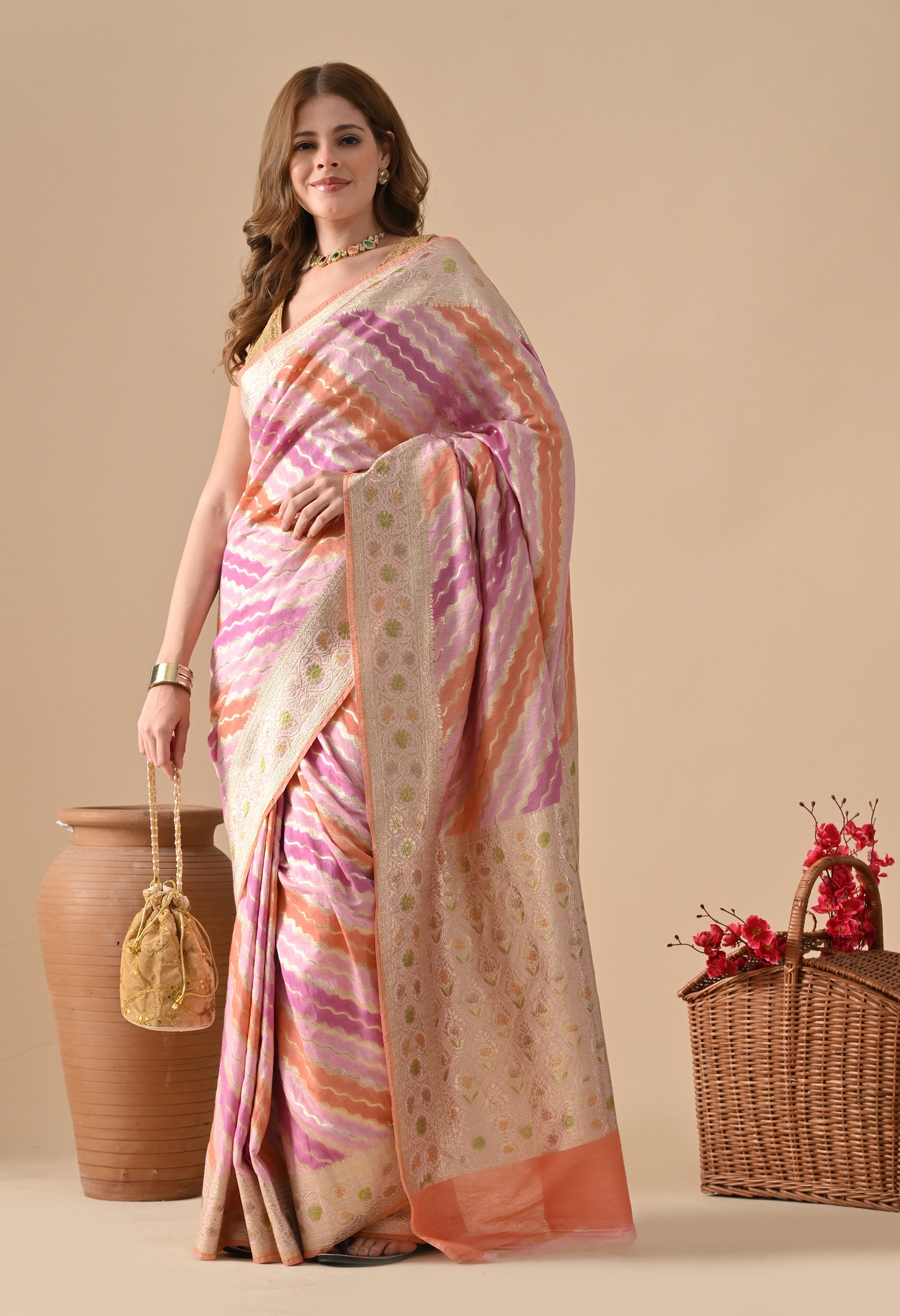 A full-length front view of a model in a The Weavers Shop by Raavi pink and orange Rangkat Banarasi saree. She is styled for a wedding reception, holding a matching gold embroidered potli bag. The image clearly shows the wavy pattern of the silk fabric and the intricate gold floral border, set against a neutral studio background with rustic props.