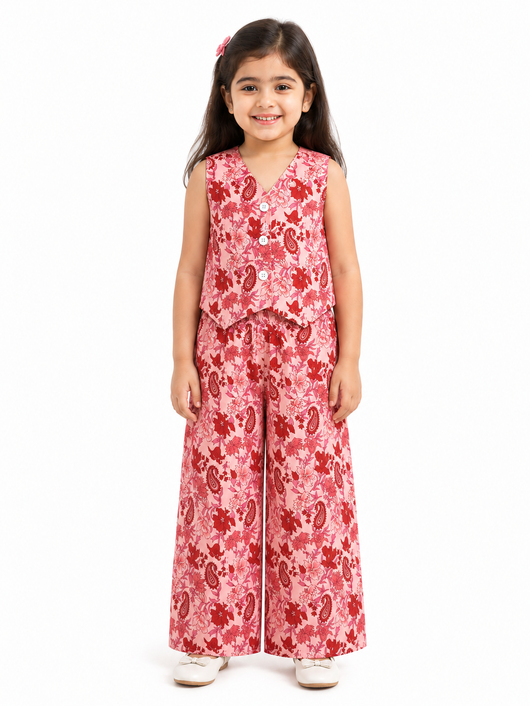 A young girl smiling in a pink pure cotton co-ord set featuring a red floral and paisley print, consisting of a sleeveless vest-style buttoned top and wide-leg trousers
