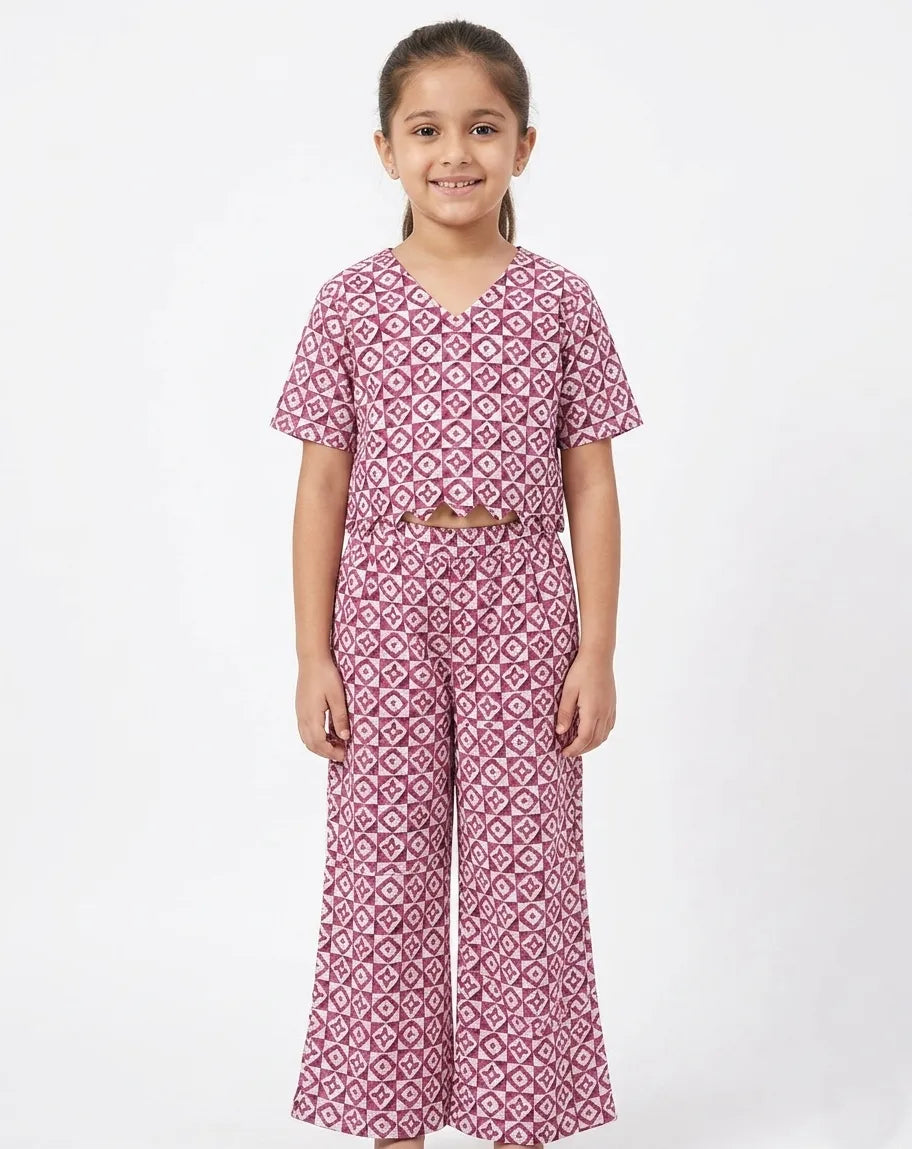 A young girl modeling a mauve and white geometric print ethnic co-ord set. The ensemble features a V-neck, short-sleeved crop top with a scalloped hem and matching high-waisted wide-leg trousers, showcasing an all-over intricate patterned design on a white background.