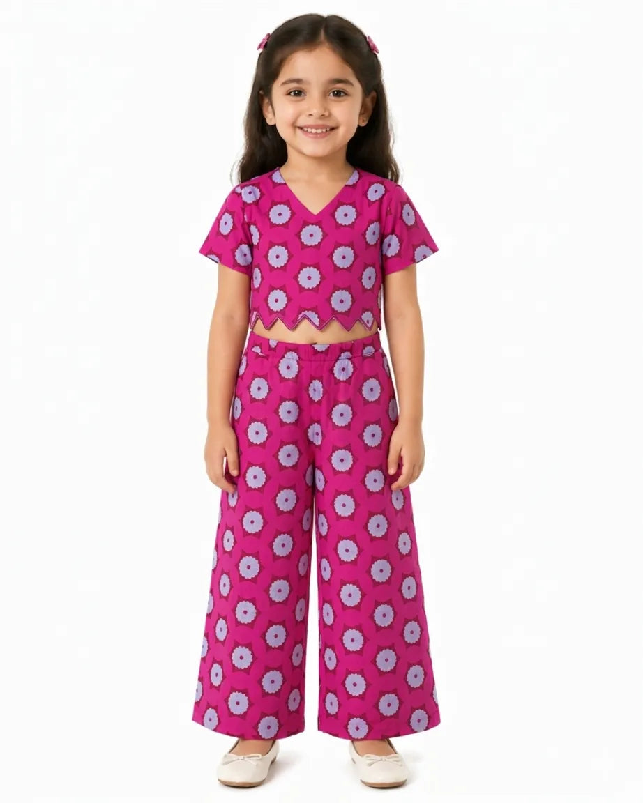 A young girl modeling a vibrant pink ethnic co-ord set featuring a short-sleeved crop top with a scalloped hem and matching wide-leg trousers. The outfit is adorned with a repeating circular floral print in purple and white.