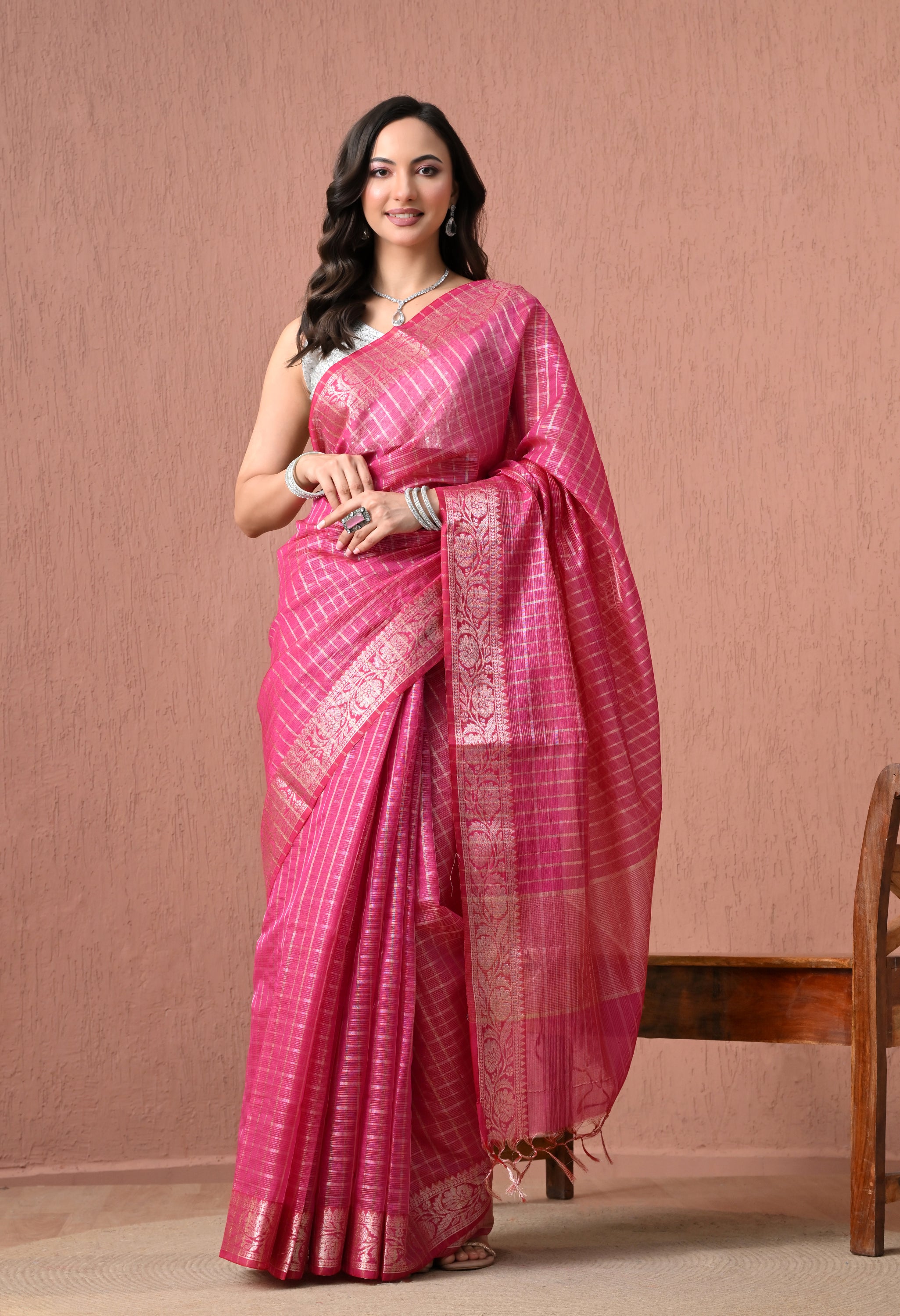 A full-length studio photograph of a woman smiling while modeling a vibrant rani pink Chanderi silk-blend saree. The saree is adorned with an all-over fine metallic gold grid (zari checks) pattern and features a wide, intricately woven gold zari border with detailed floral motifs.
